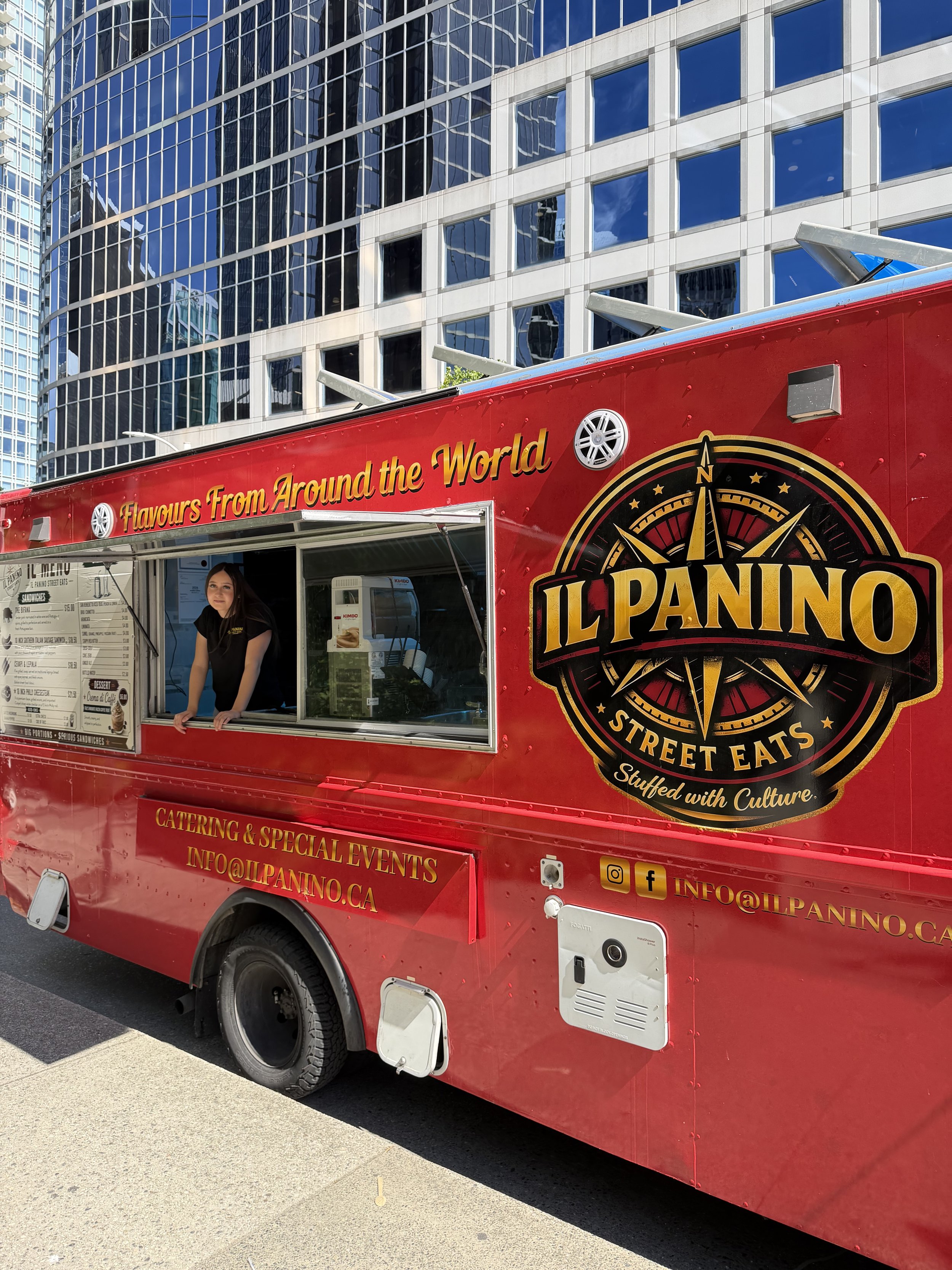 A red street food truck named Il Panino, offering flavors from around the world, parked in an urban area with tall glass buildings in the background. The truck features a large logo with a compass and the slogan 'Stuffed with Culture.' A woman is standing inside the serving window.