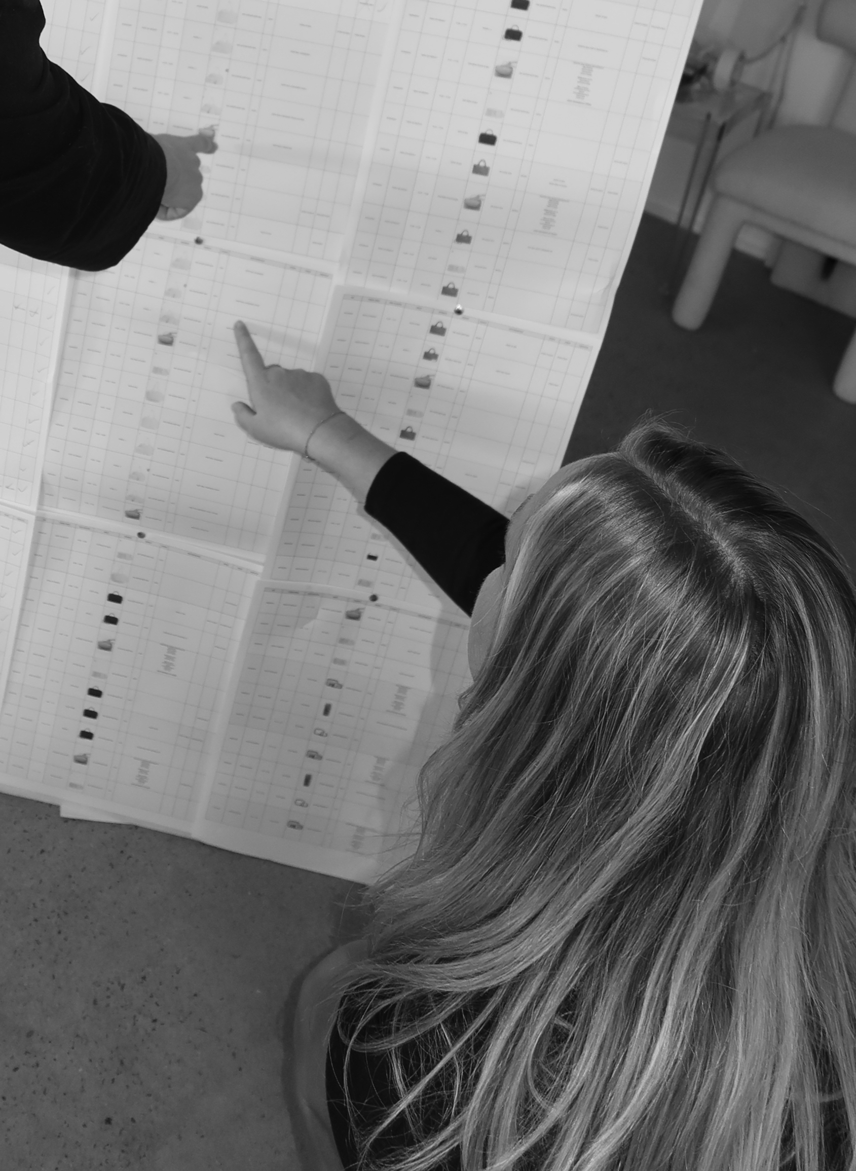 A person with long hair is pointing at a large printed chart on a table, which appears to contain multiple rows and columns of data and images. The photo is in black and white and shows a part of a room with a chair or couch in the background.