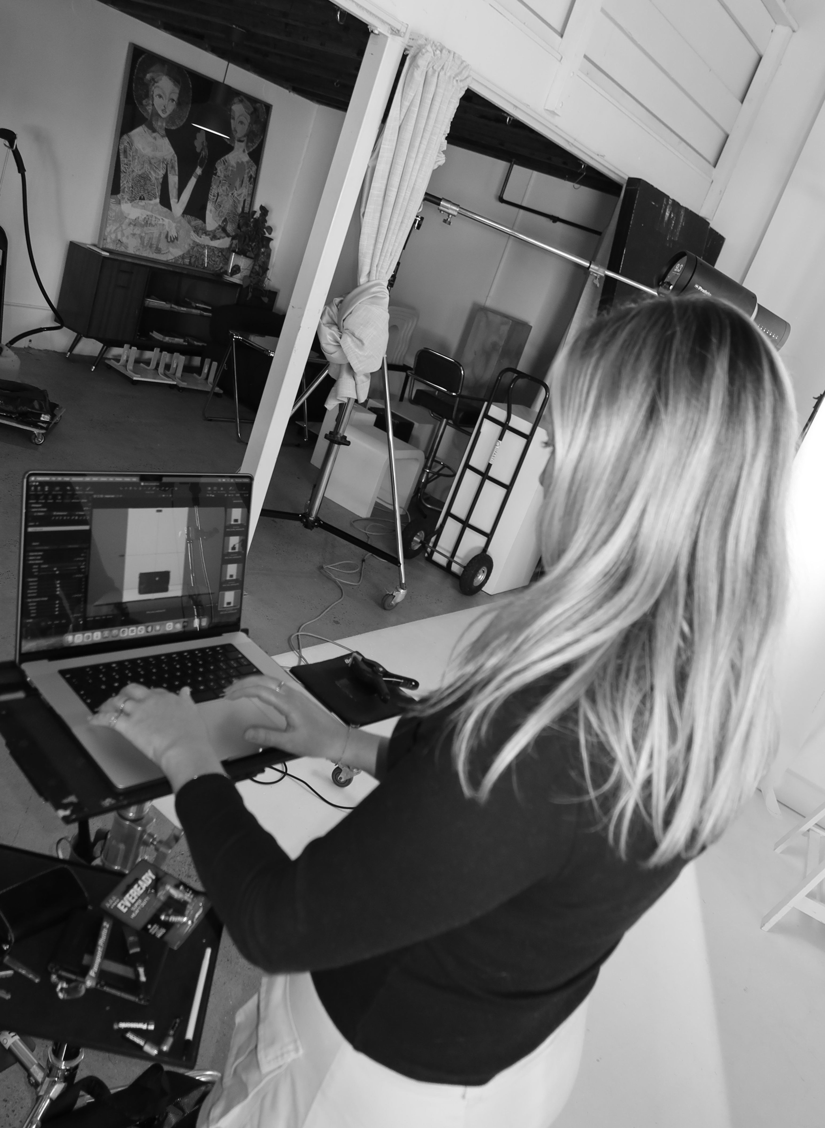 A person with long blonde hair working on a laptop at a desk in a room with photoshoot equipment, items on the desk, and a wall with artwork.