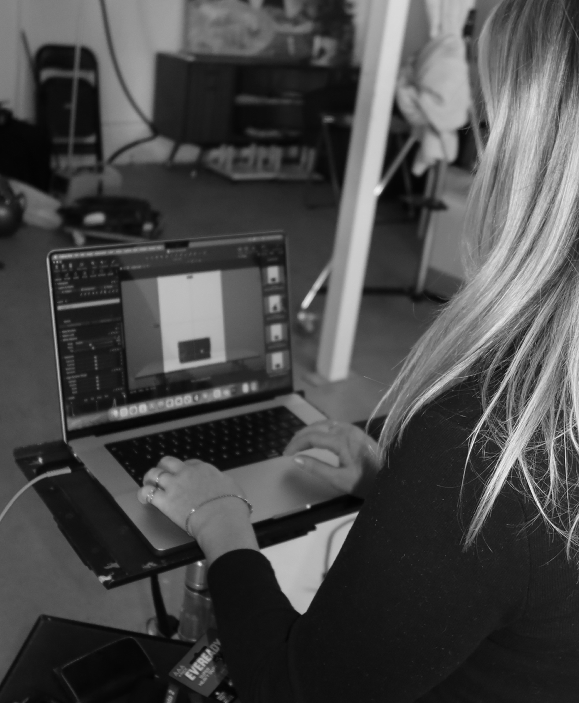 A person with long hair sitting at a table working on a laptop computer in a room with various furniture and objects in the background.