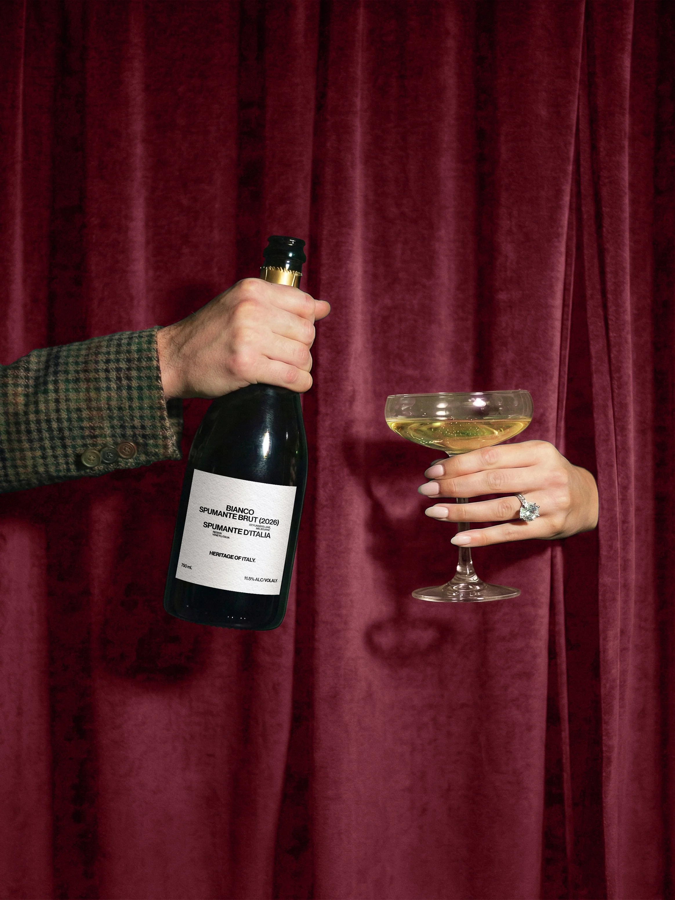 Person holding a bottle of Italian sparkling wine and a glass of white wine against a red velvet curtain.