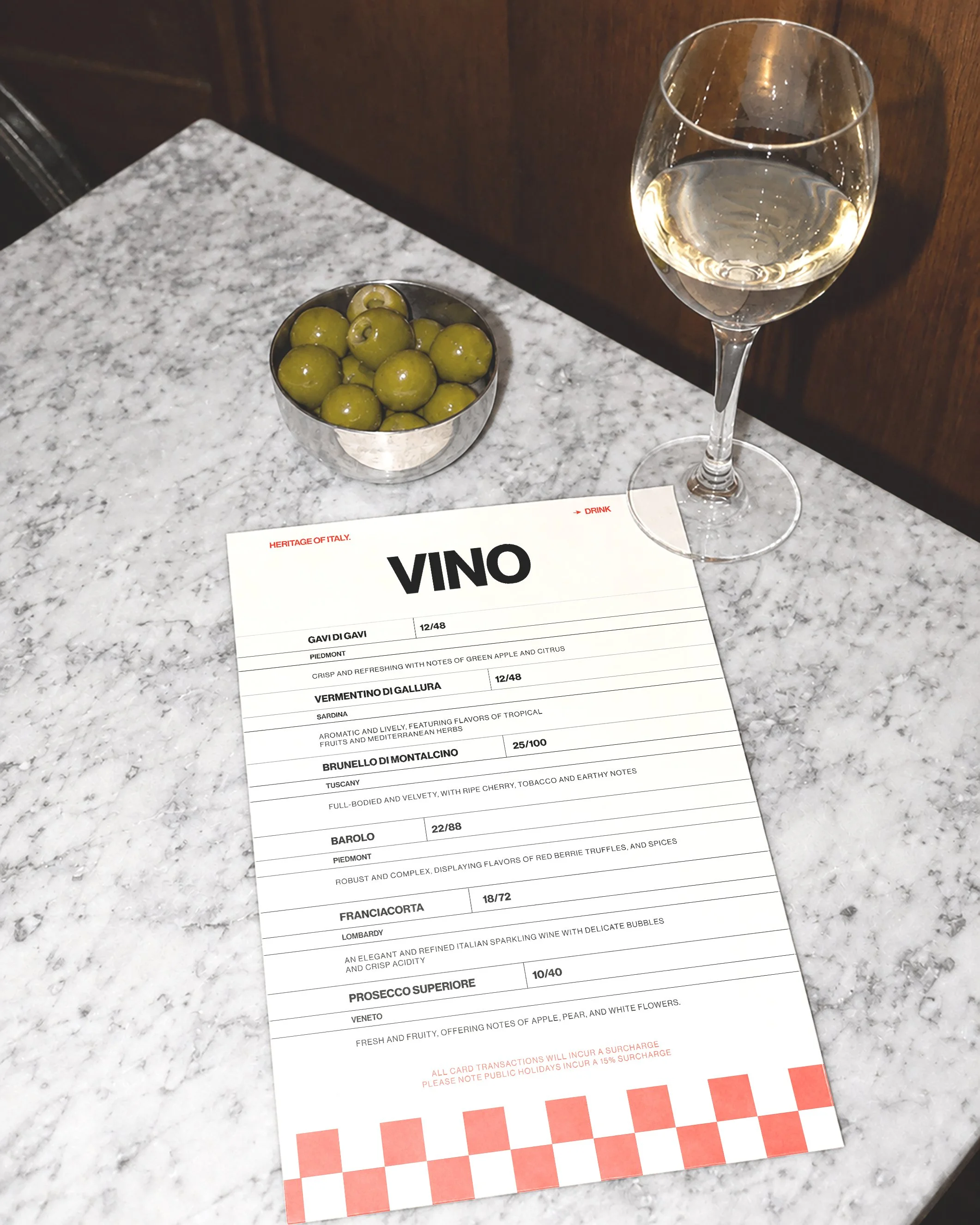 A wine tasting menu, a glass of white wine, and a small bowl of green olives on a marble table.