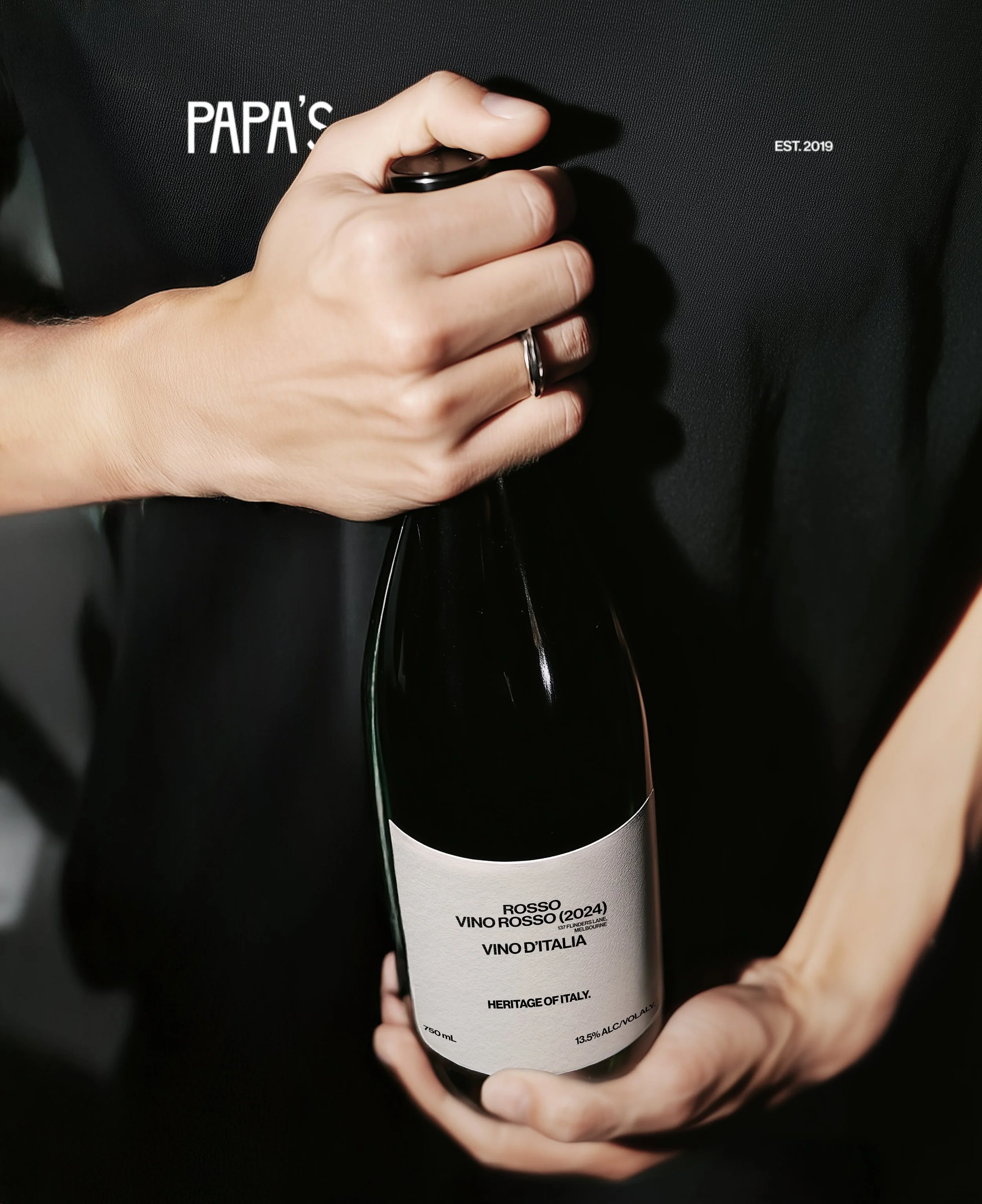 Person holding a wine bottle labeled 'Rosso Vino Rosso 2024, Heritage of Italy, 13.5% alcohol, 750 ml, 137 Flinders Lane, Melbourne, Australia' in front of a dark background.