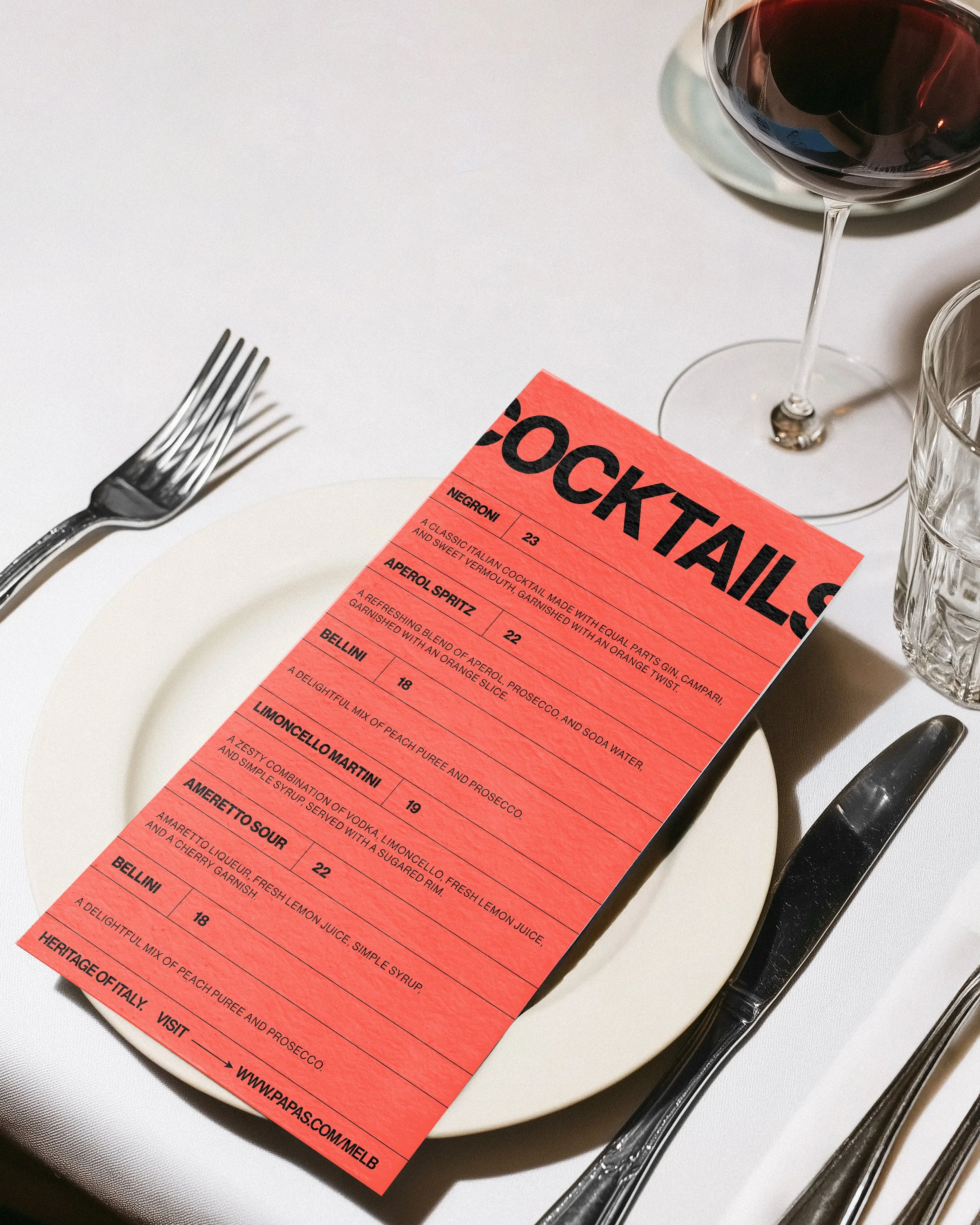 A table setting with a fork, a glass of red wine, and a menu on a white plate and a glass of water.