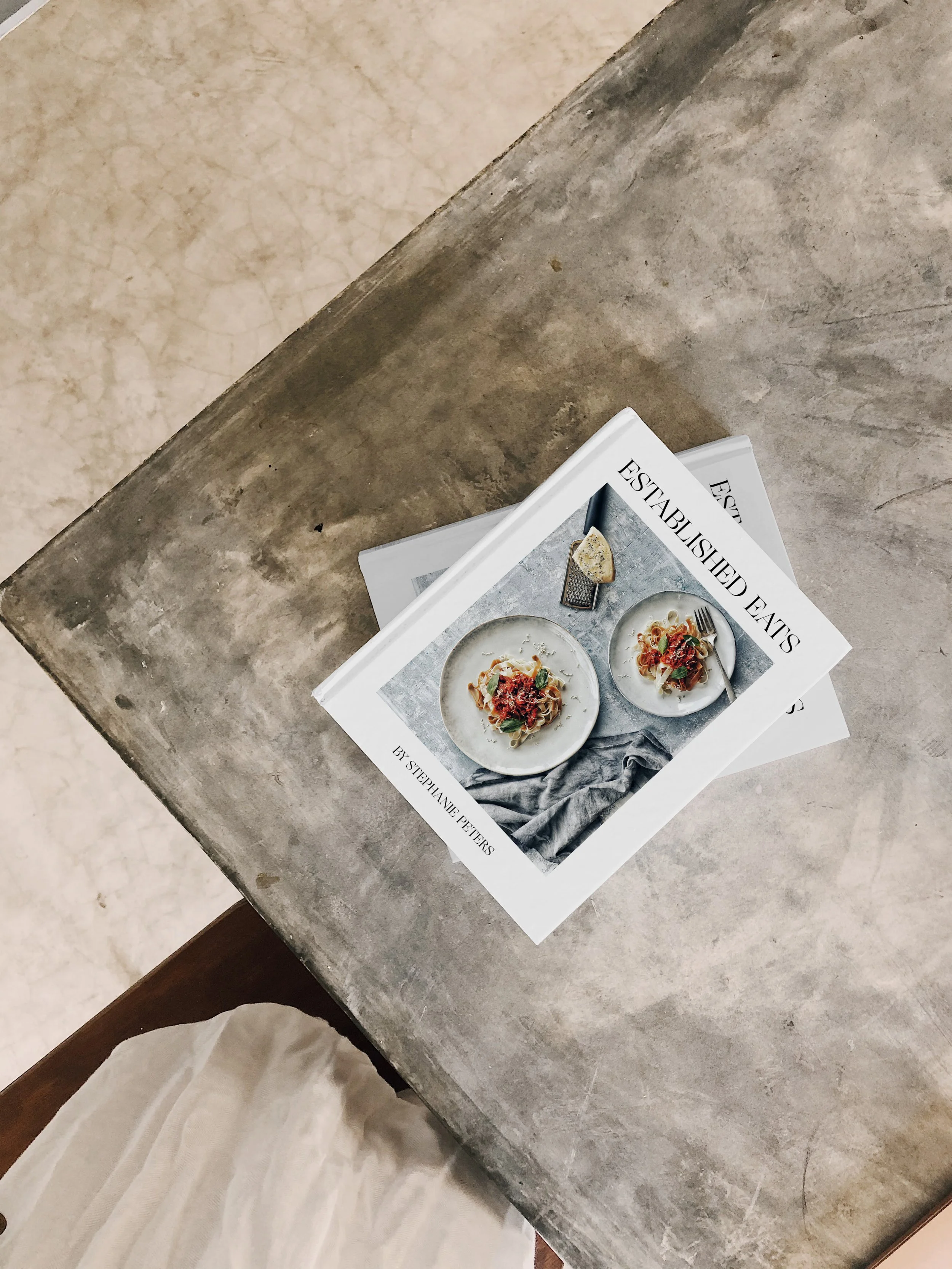 A cookbook titled 'Established EATS' featuring images of plates of pasta dishes rests on a gray concrete surface, with a textured beige surface partially visible nearby.