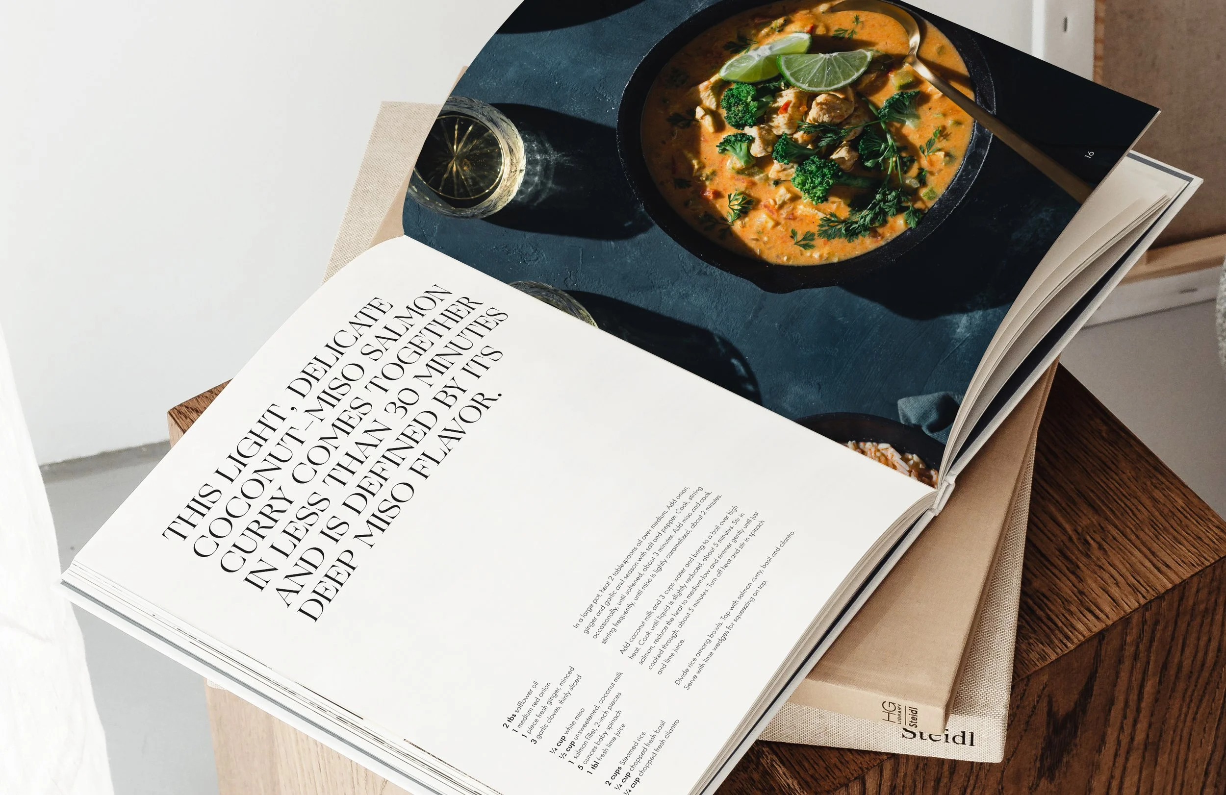 An open cookbook on a table displaying a recipe page and a photograph of chicken curry garnished with cilantro, lime wedges, and vegetables, with a glass of lime-infused water beside it.