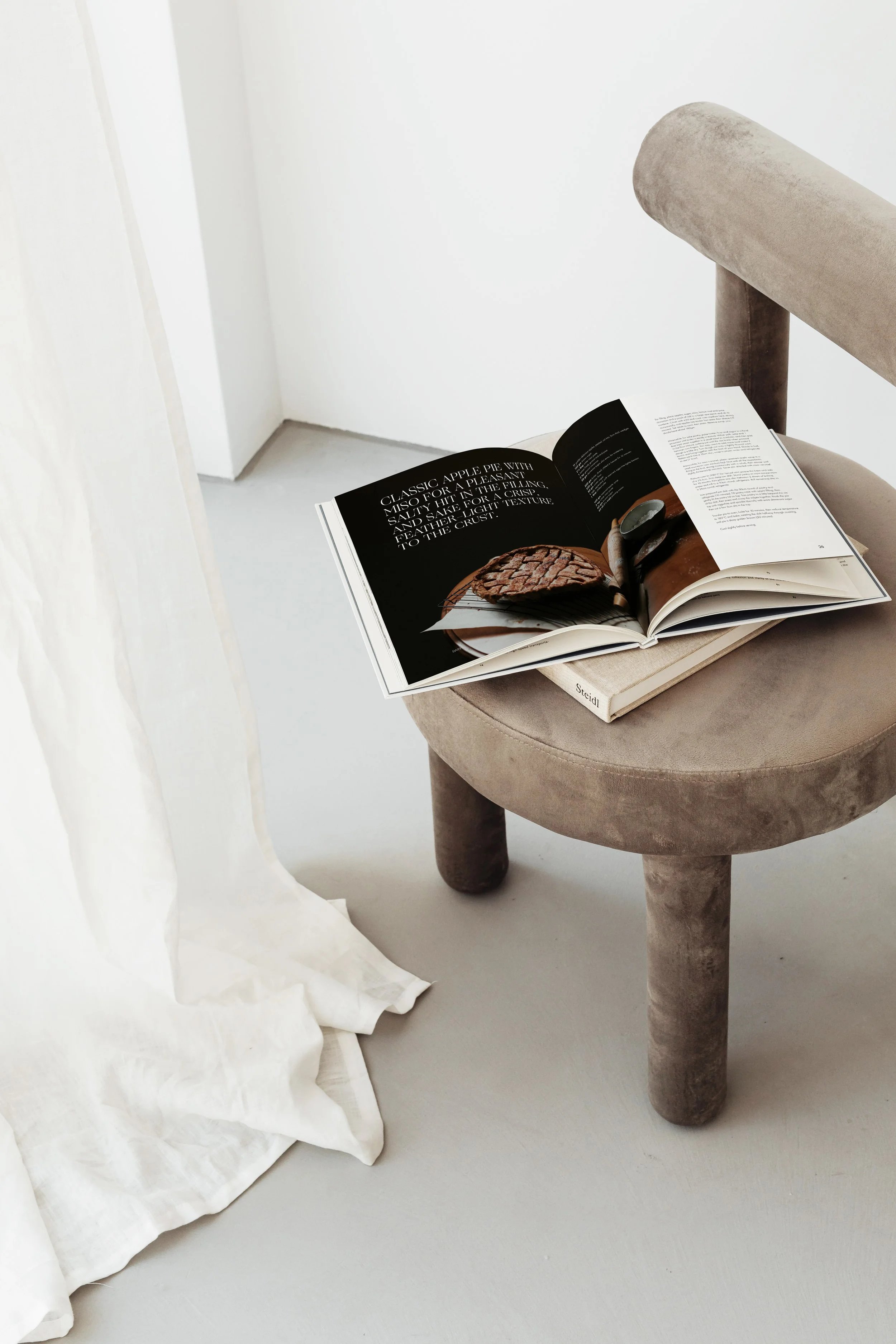 An open magazine on a beige upholstered stool with a wooden leg, next to white flowing curtains, on a white floor and wall background.