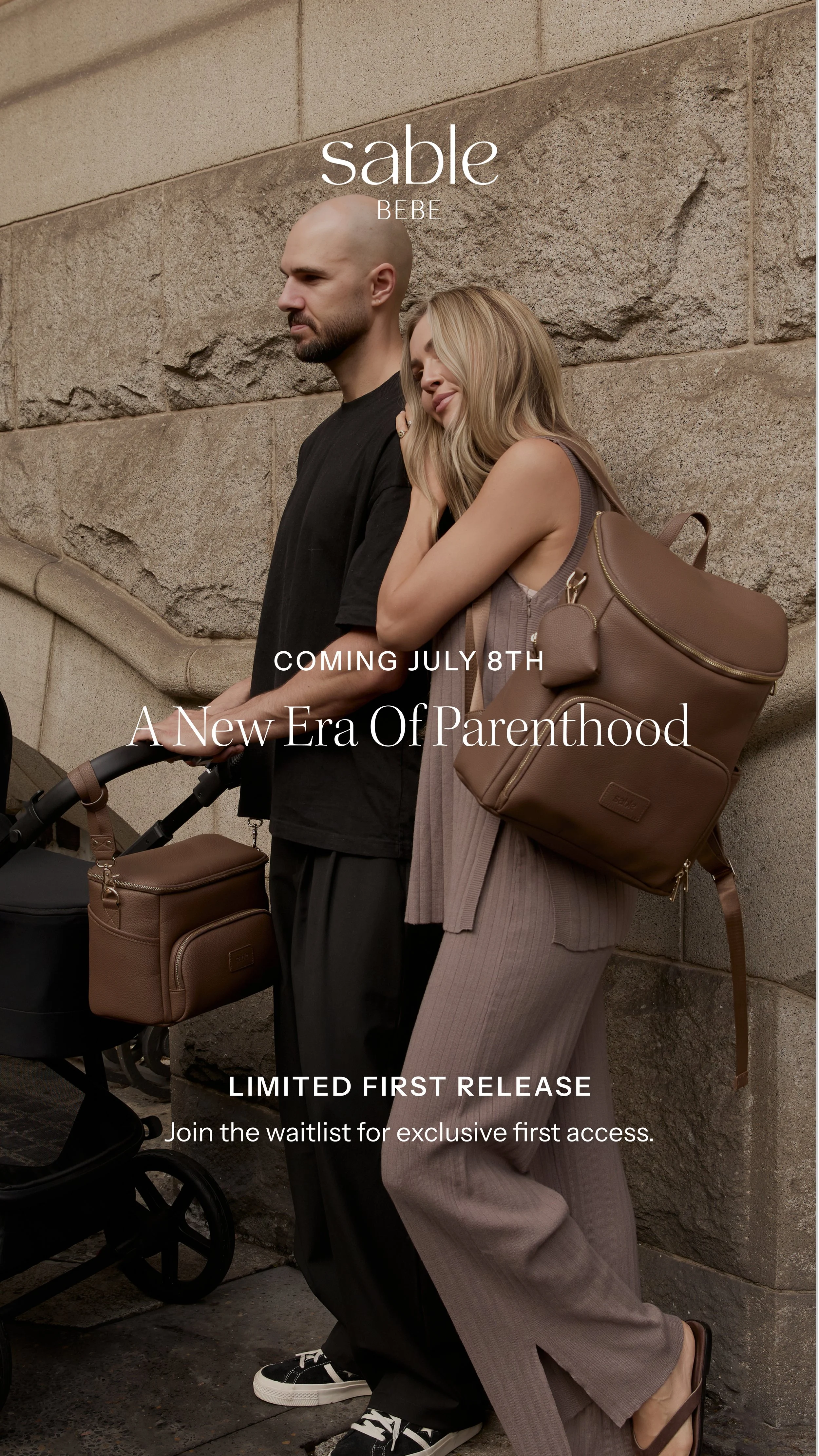 Two people, a man and a woman, leaning against a stone wall, with the woman resting her head on the man's shoulder. They are dressed in casual clothing and carrying matching brown bags. A stroller with a matching brown bag is partially visible in fro