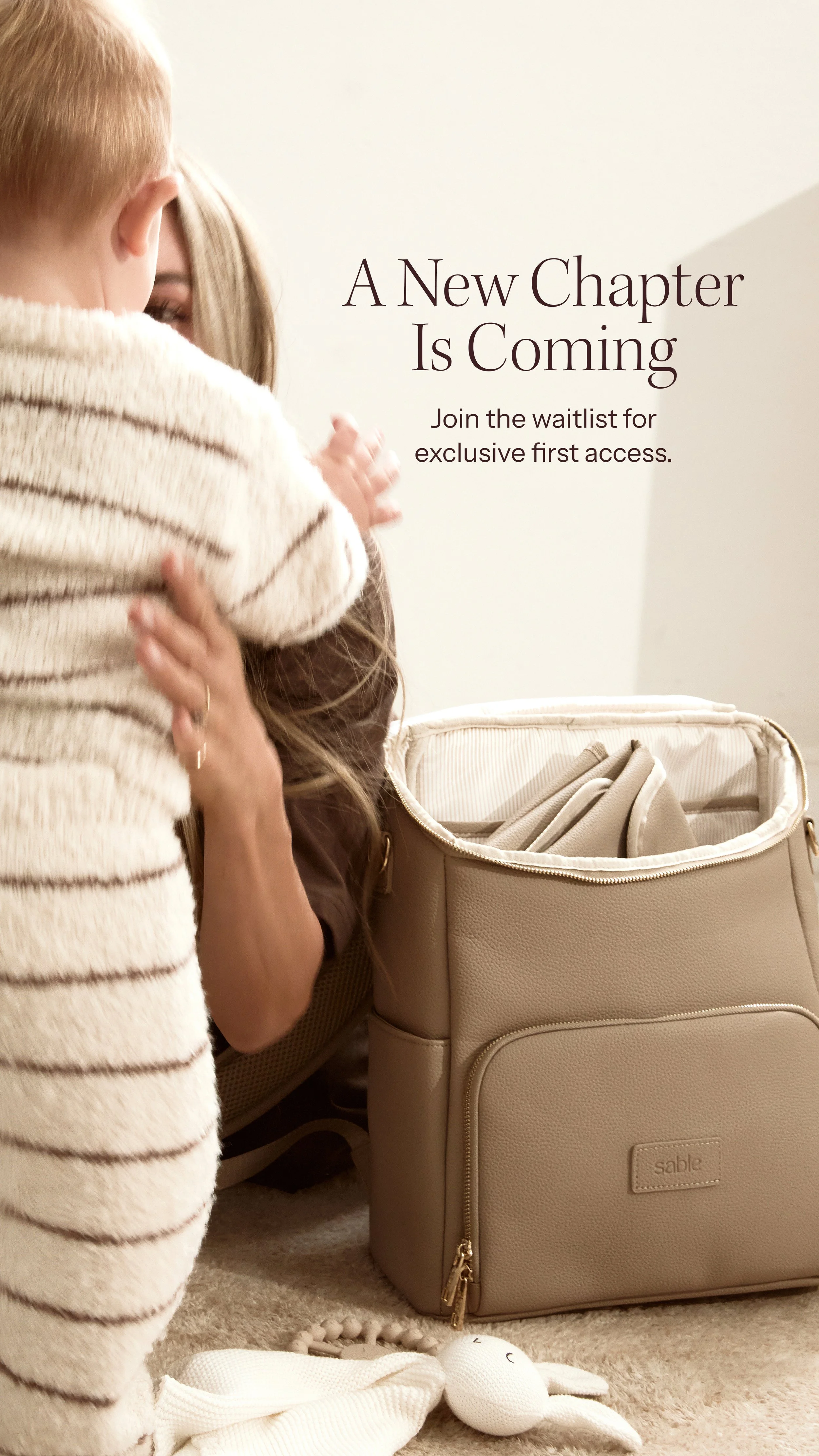 A woman is holding a young child in her arms, near a beige diaper bag that has its zipper open. The diaper bag contains some items, and there is a plush bunny toy with a smiling face on the beige carpet in front of them. The scene suggests preparatio