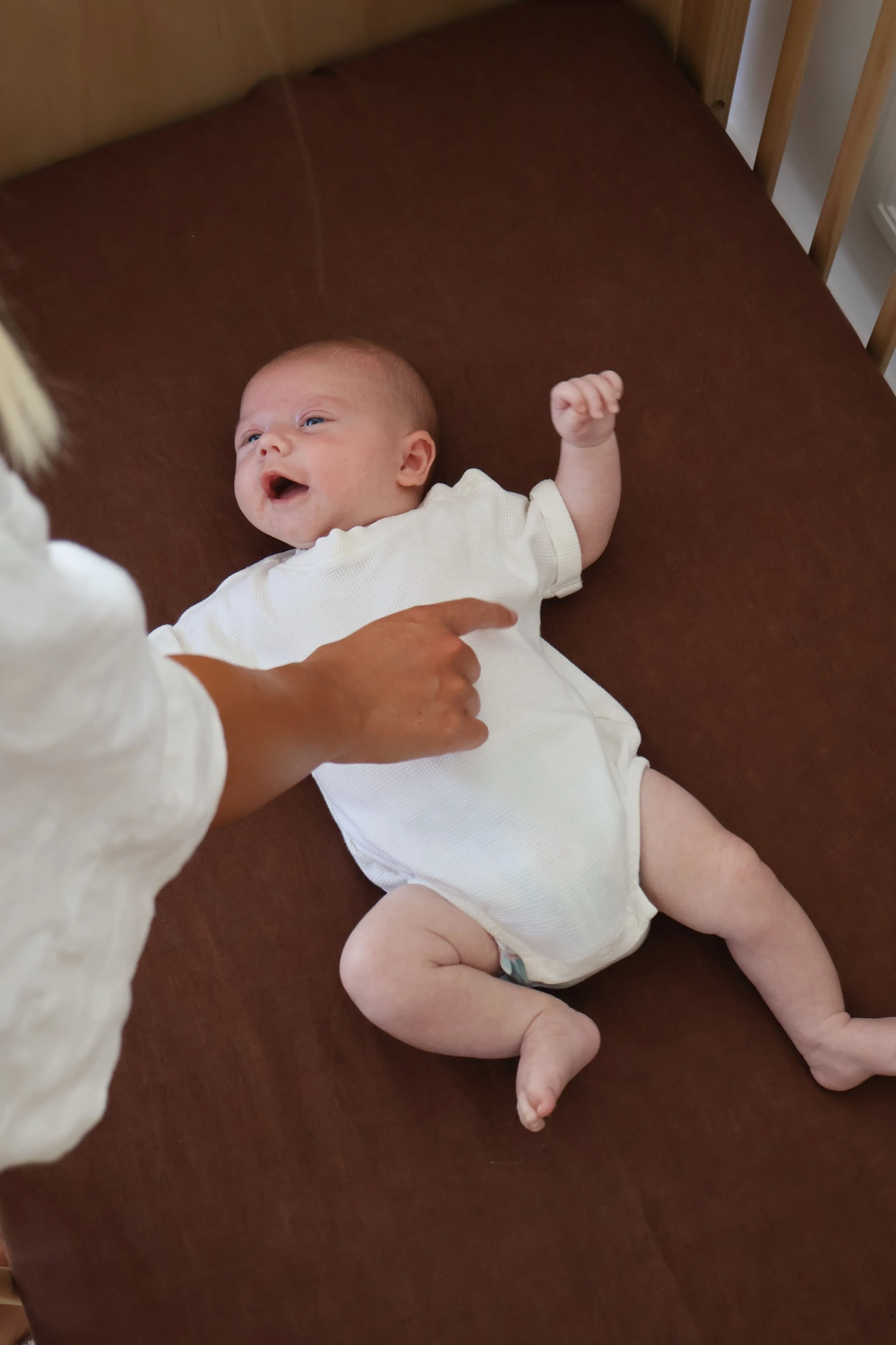A baby lying on a brown changing table, smiling and looking up, wearing a white shirt and diaper, with an adult's hand gently touching the baby's side.