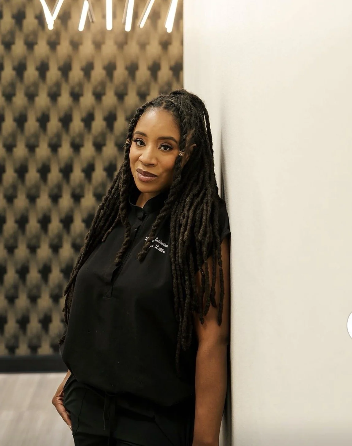 A woman with long dreadlocks leaning against a white wall, wearing black clothing, in a room with acoustic foam panels in the background.