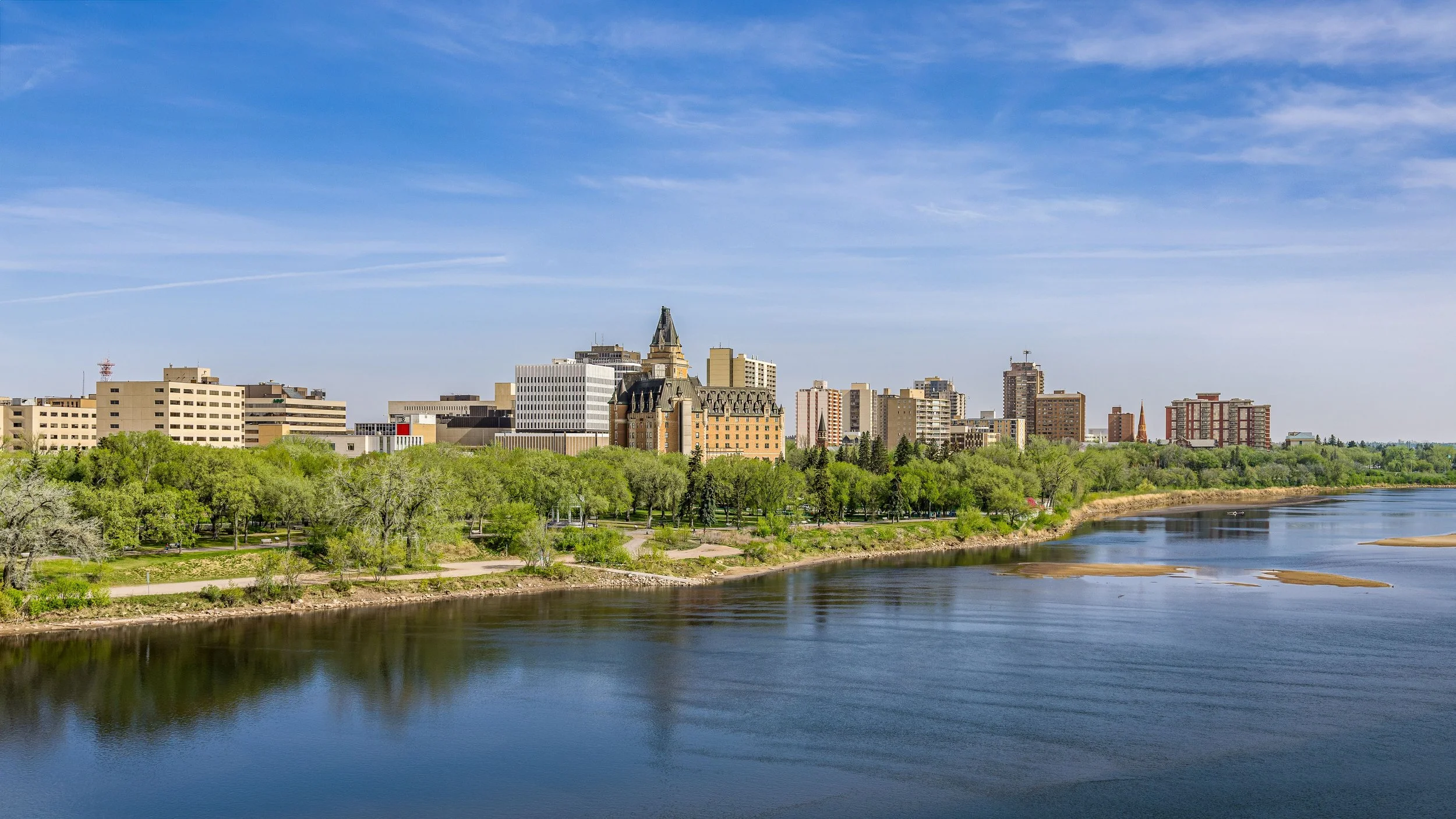 City skyline with various buildings along a riverbank, lush green trees in the foreground, and a blue sky with wispy clouds above.