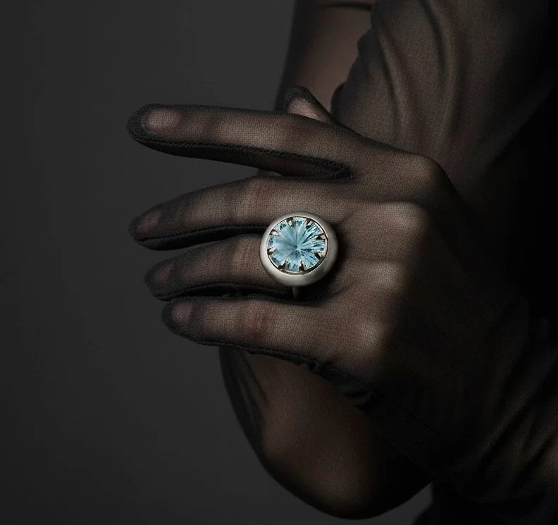 A hand wearing black gloves, showing a large ring with a blue gemstone, close-up shot against a dark background.