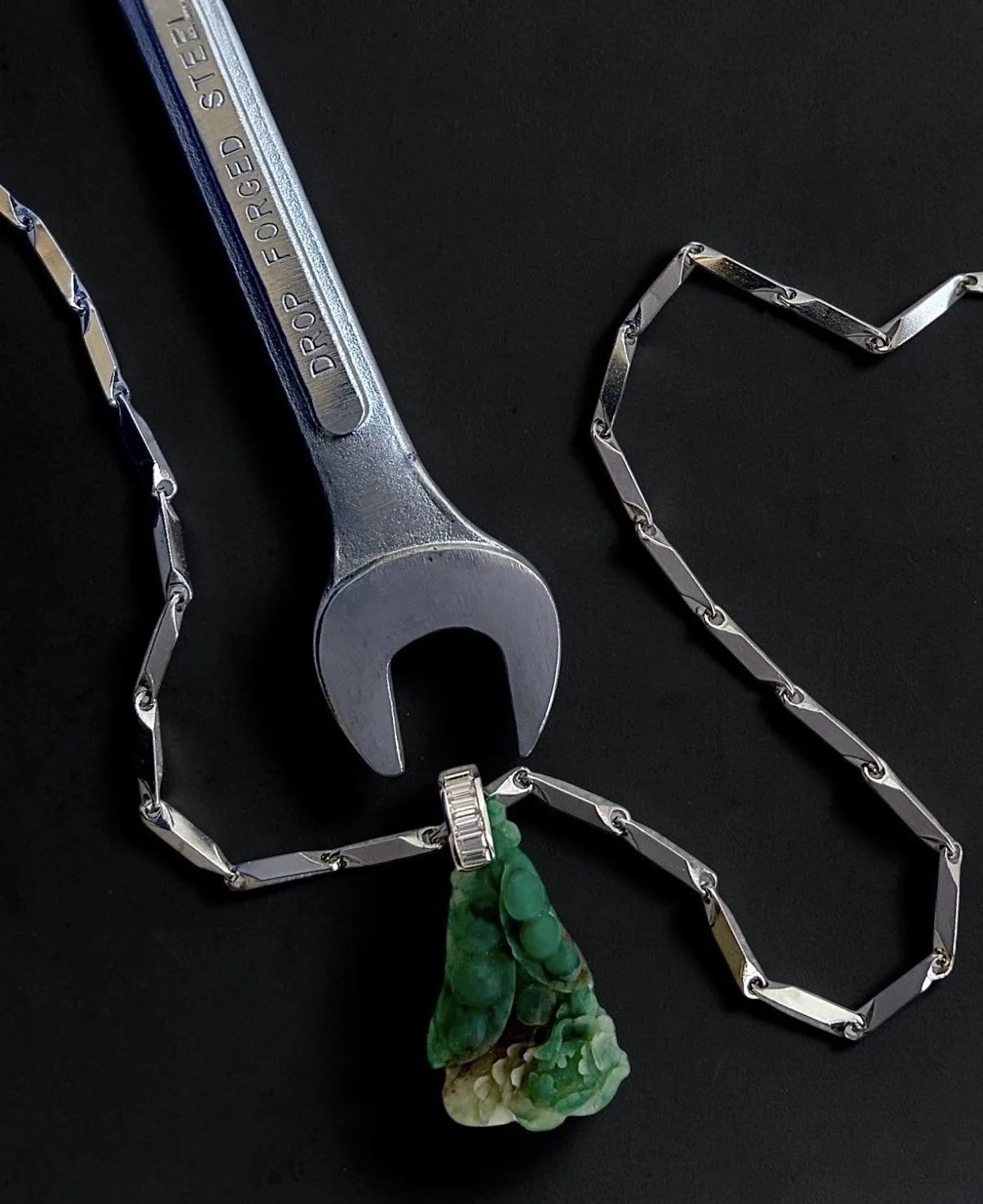 A wrench labeled 'DROP FORGED STEEL' with a chain attached to its opening, and a green and white stone pendant hanging from the chain, all on a black surface.