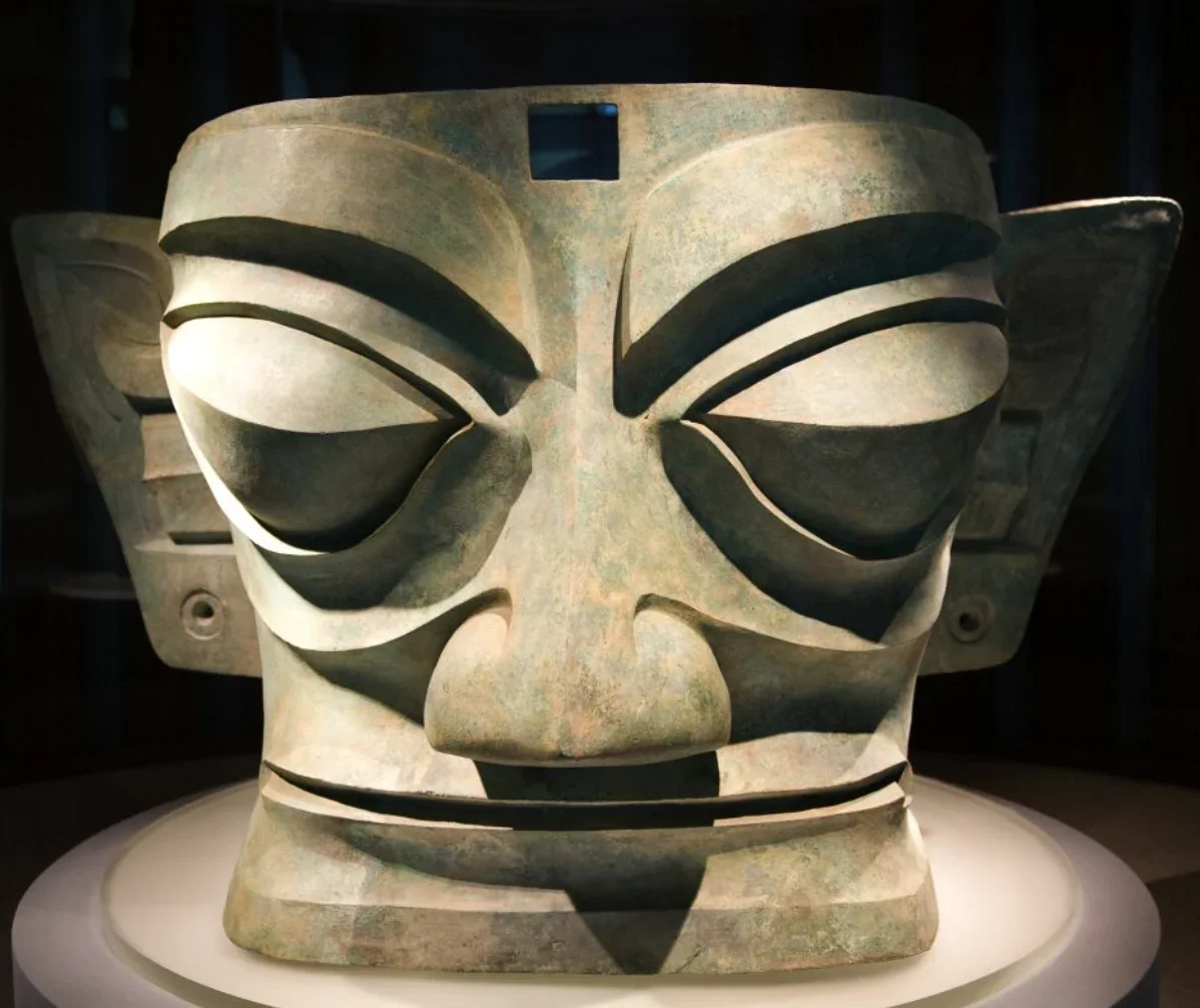 Large ancient sculpture of a human face with prominent eyebrows, nose, and mouth, carved from stone, displayed in a museum.