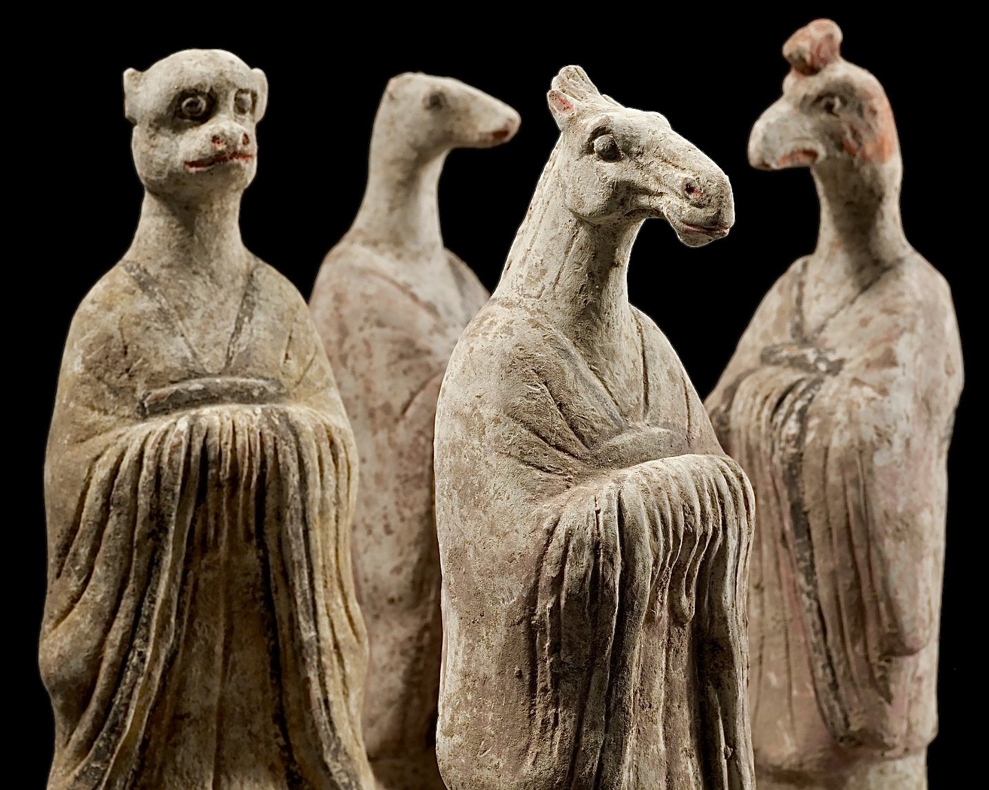 Four ancient terracotta figurines with elongated bodies and animal features, set against a black background.