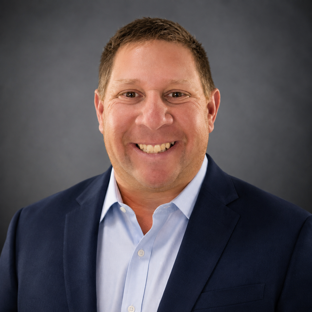 Professional portrait of a smiling man in a navy blazer and white shirt against a dark gray background.