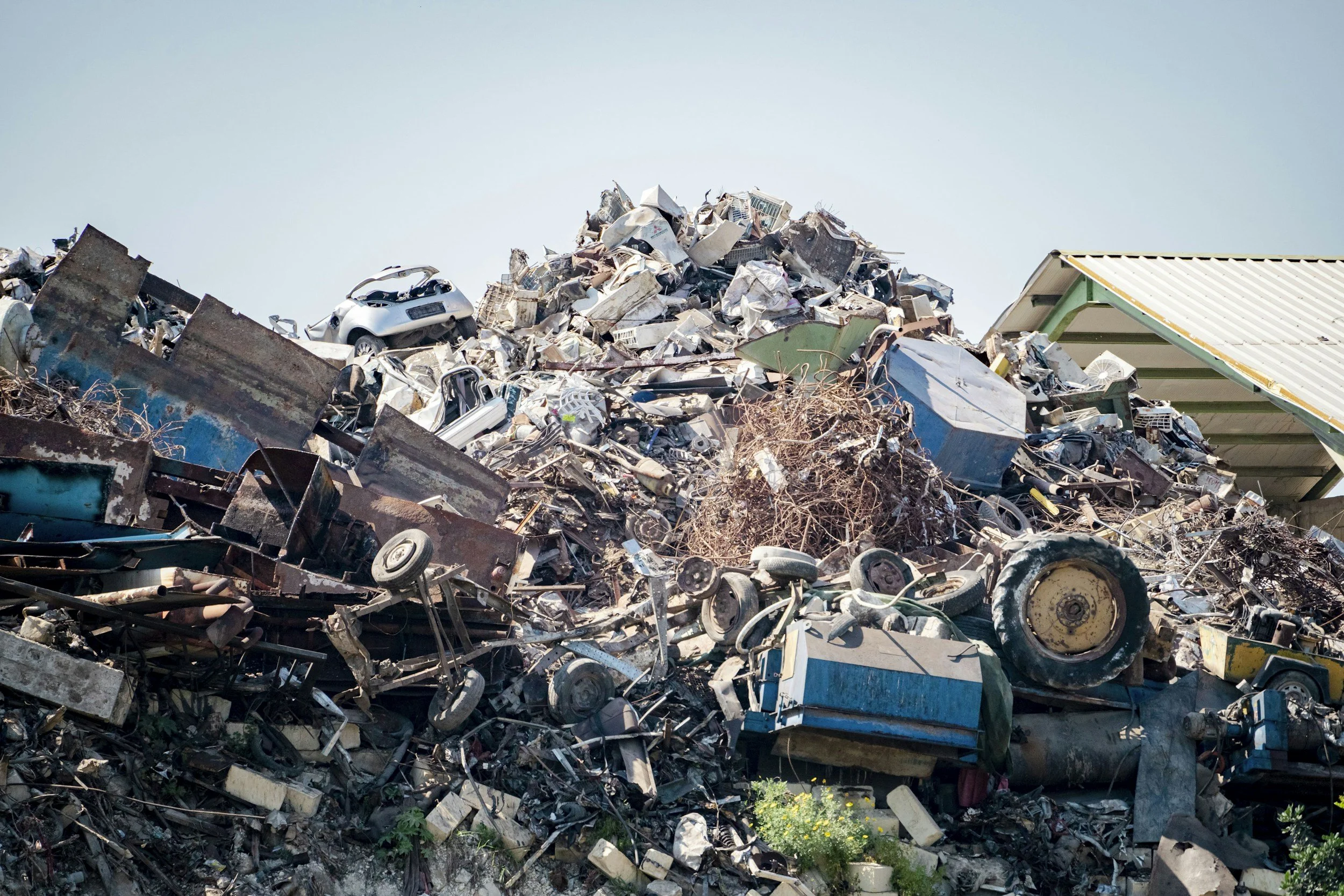 A large pile of scrap metal, car parts, and debris with some plants growing at the bottom, and a clear blue sky above.