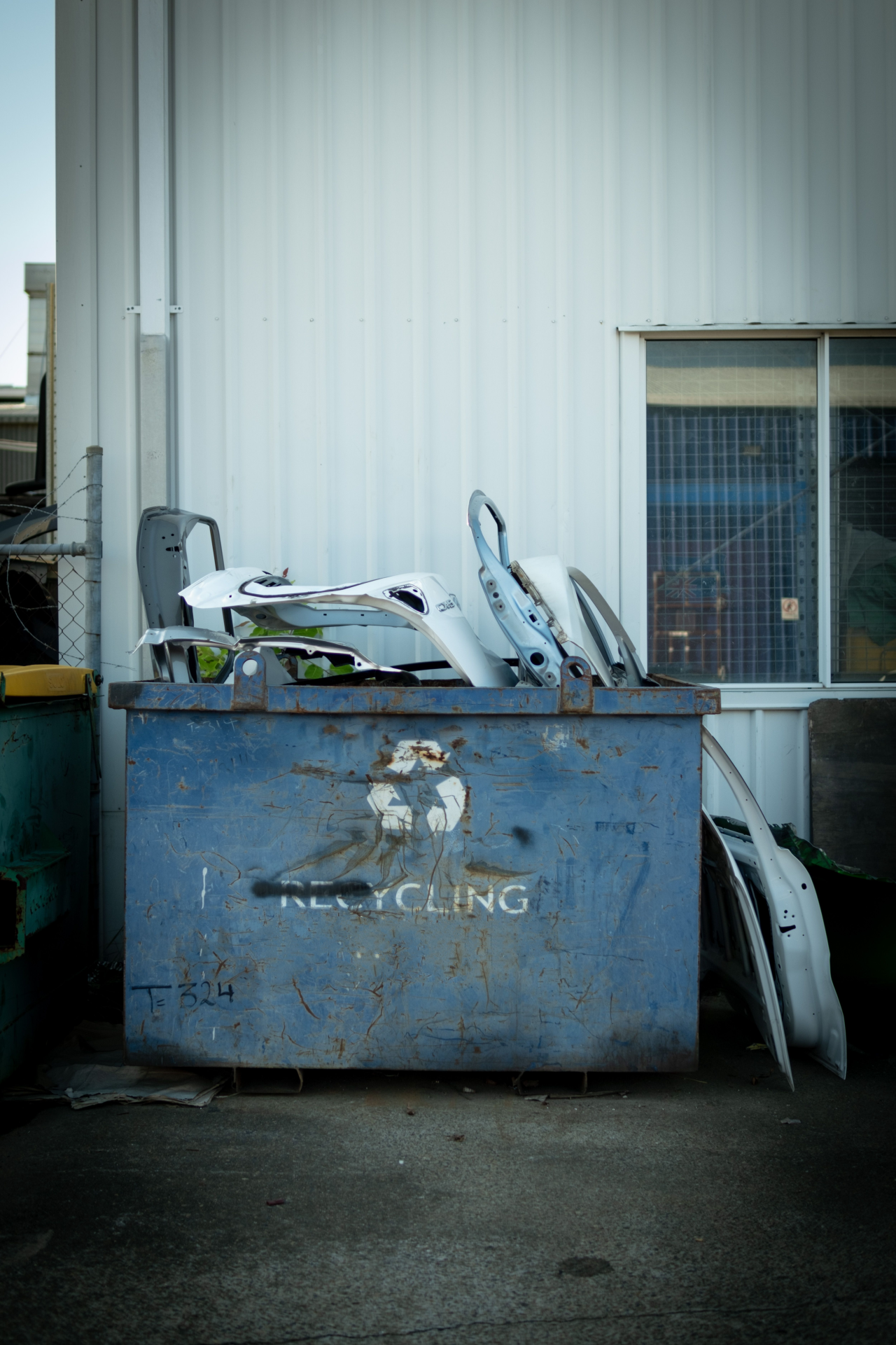 A rusty blue recycling bin filled with discarded white car parts, placed outdoors against a white metal building with a window.