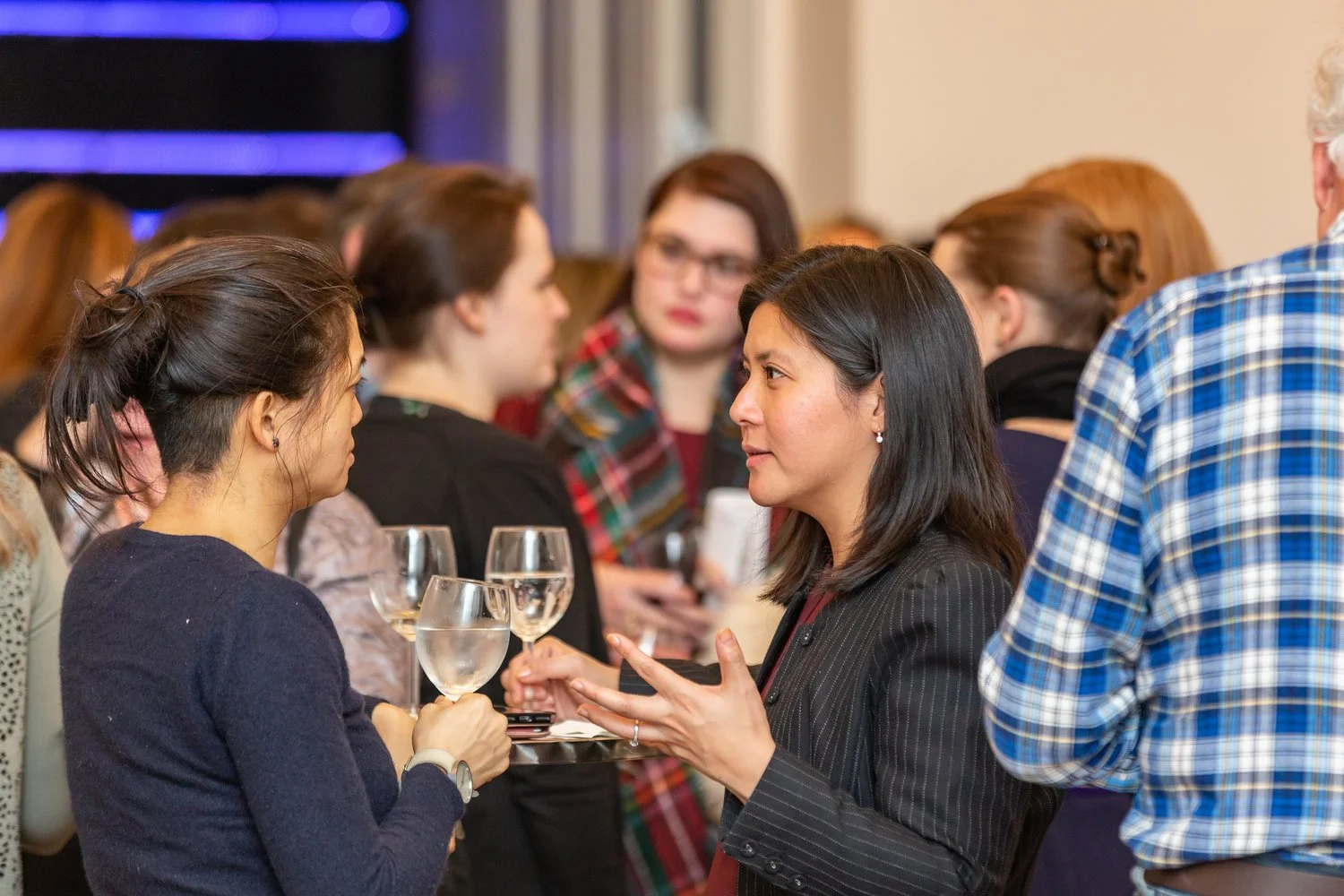 Two women converse at a social gathering with glasses of wine, while other people mingle in the background.