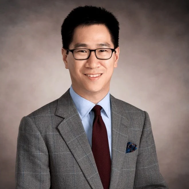 Professional headshot of a young man with black hair, glasses, in a gray plaid suit, light blue shirt, maroon tie, and pocket square, smiling against a neutral background.