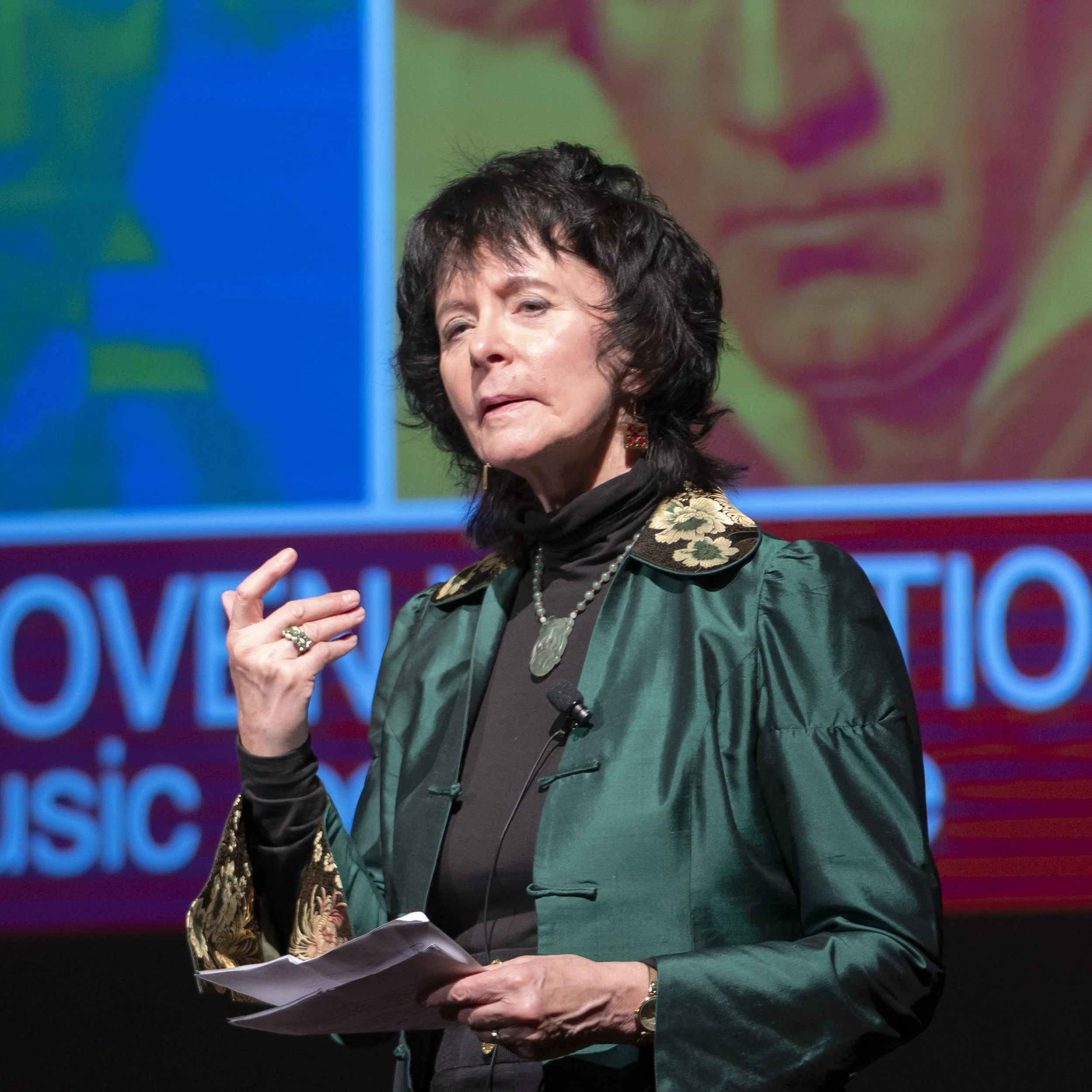 Close-up of a woman with dark, curly hair speaking at an event, holding notes, wearing a green pinstripe jacket and earrings, with a colorful background featuring blue and green hues.