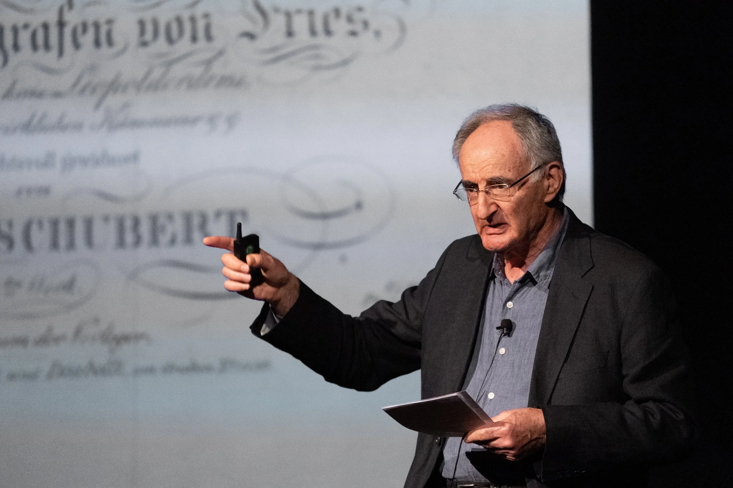 A man with glasses, gray hair, and a serious expression, wearing a dark blazer and striped shirt, giving a presentation or speech with a remote control in his hand. A large screen behind him displays blurred text.