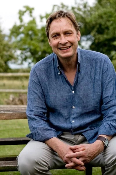 A man sitting on a bench outdoors, smiling, wearing a blue button-up shirt and beige pants, with trees and a wooden fence in the background.