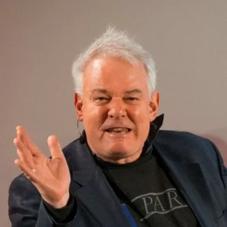 A man with white hair wearing a dark blazer and black T-shirt, gesturing with his right hand, smiling slightly against a neutral background.