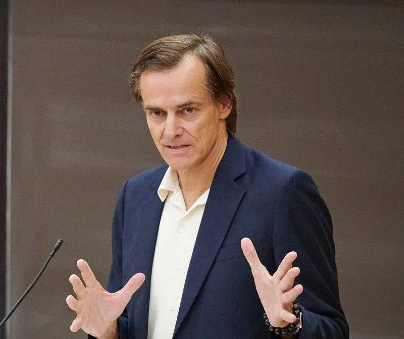 A middle-aged man with brown hair, wearing a dark blue blazer and a white shirt, gesturing with his hands while speaking or presenting.