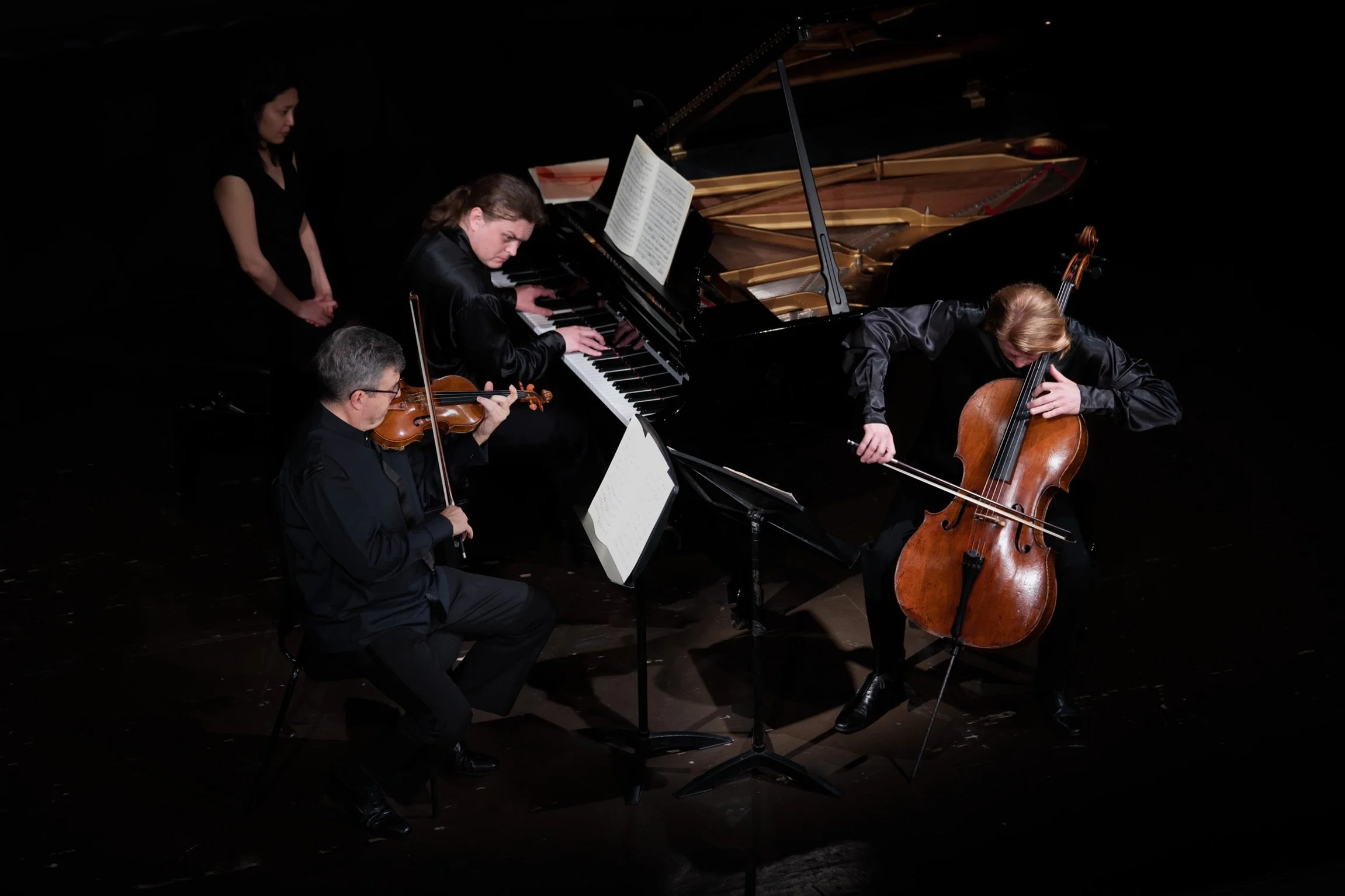 Four musicians performing with a grand piano, violin, and cello on stage in a dark setting.