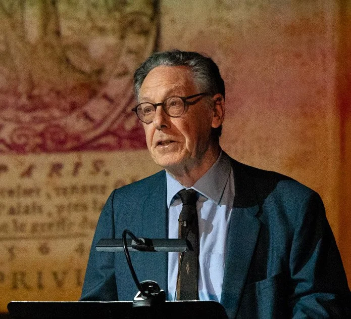 An older man in a blue suit and glasses speaking at a podium with a microphone, background features a blurred historical or old document.