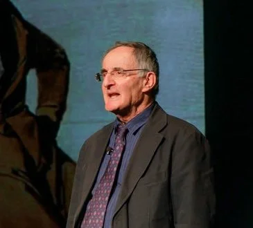 A man with glasses and gray hair is speaking or singing, wearing a gray suit, blue shirt, and patterned tie. He is on a stage with a dark background.