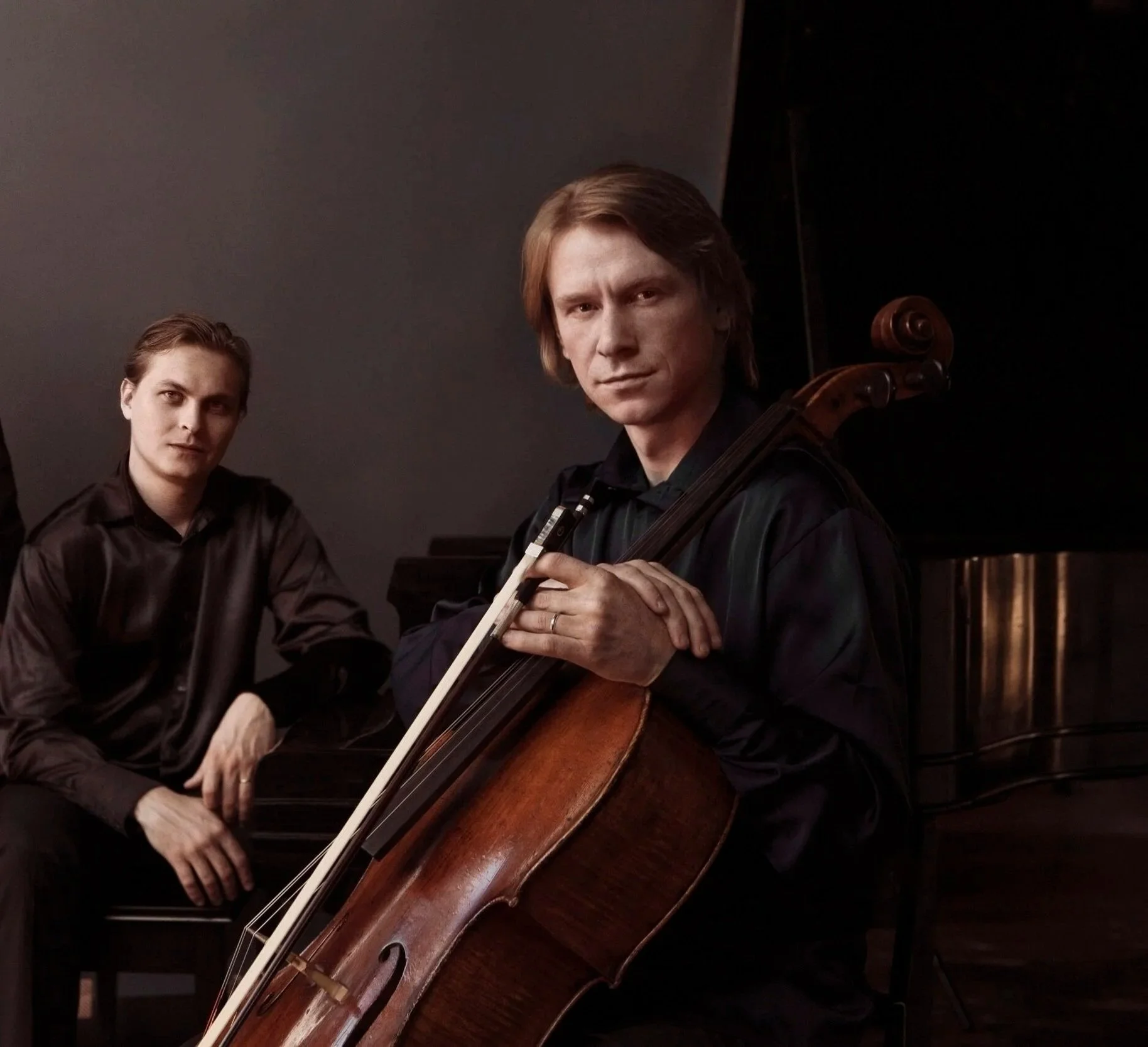 Two musicians, one playing a grand piano and the other playing a cello, in a dimly lit room.