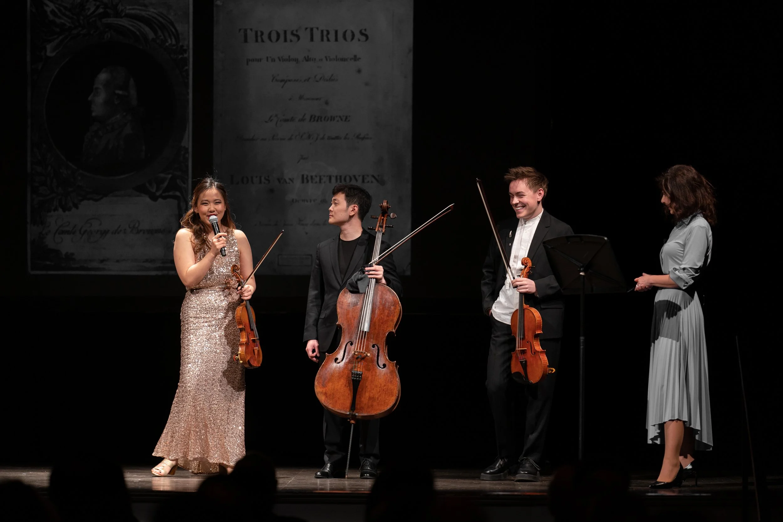 Four musicians on stage, three holding violins and one holding a cello, smiling and engaging with each other after a performance. A backdrop features a portrait and sheet music related to Beethoven's 'Trois Trios'.