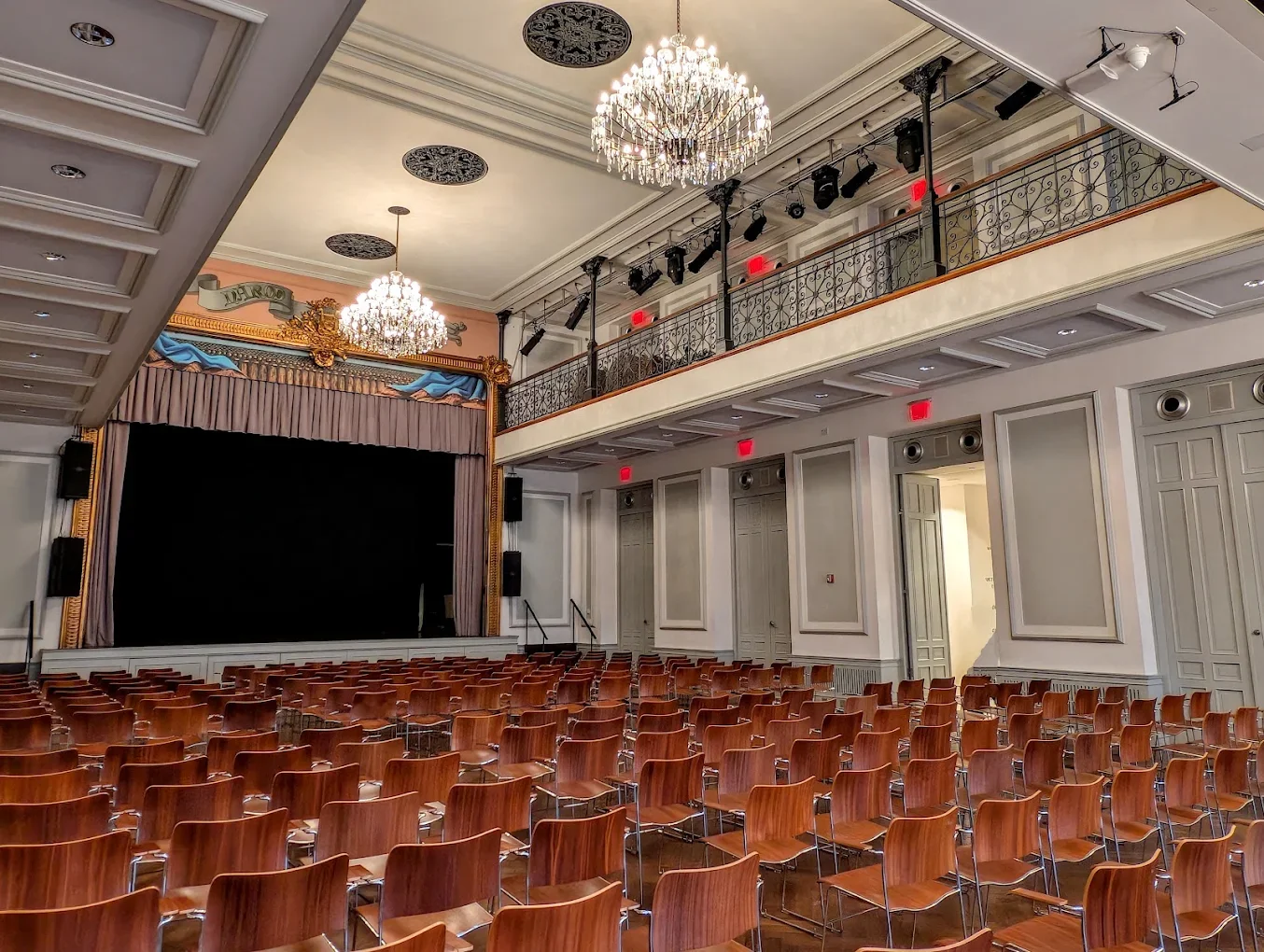An empty auditorium with rows of wooden chairs, a stage with curtains, chandeliers, and a balcony with ornate railing.