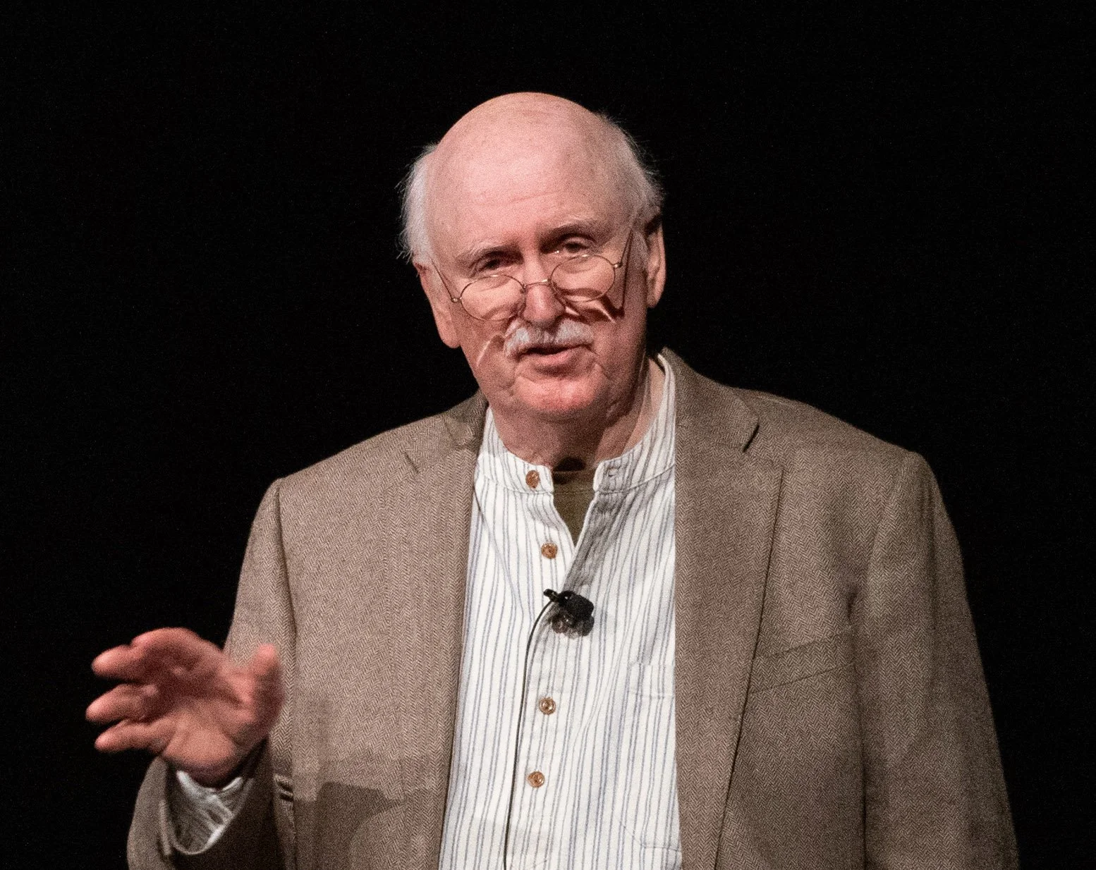 An older man with glasses and a mustache wearing a beige blazer over a striped shirt, standing against a black background, possibly giving a presentation or speech.