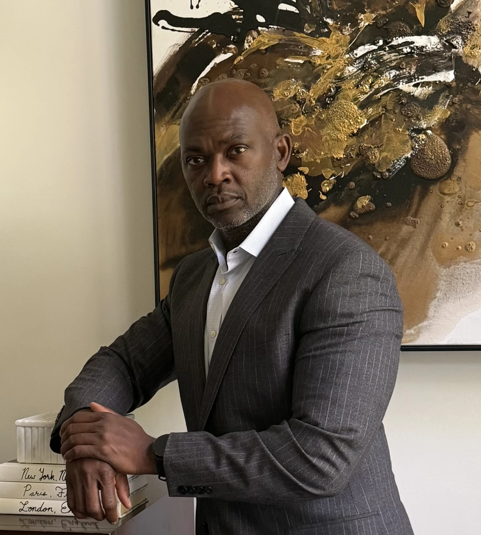 A man with dark skin and a receding hairline wearing a dark pinstripe suit and white shirt, standing indoors in front of an abstract painting with earth tones and gold accents.