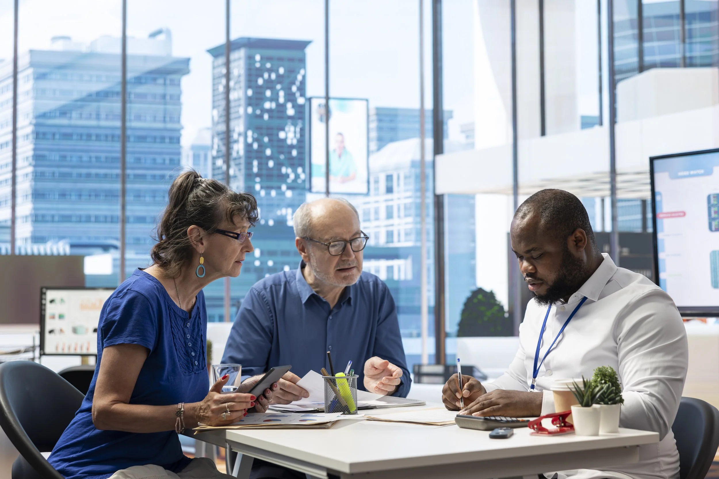 Three diverse people collaborating at a modern office table with city buildings visible through large windows in the background.
