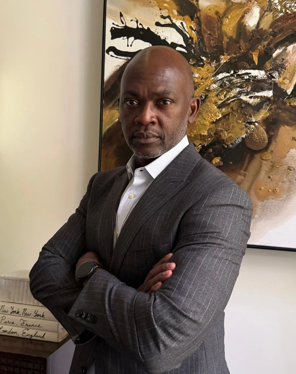 A man in a gray pinstripe suit with a white shirt standing with arms crossed in front of a large, abstract painting with swirling gold, black, and brown colors.