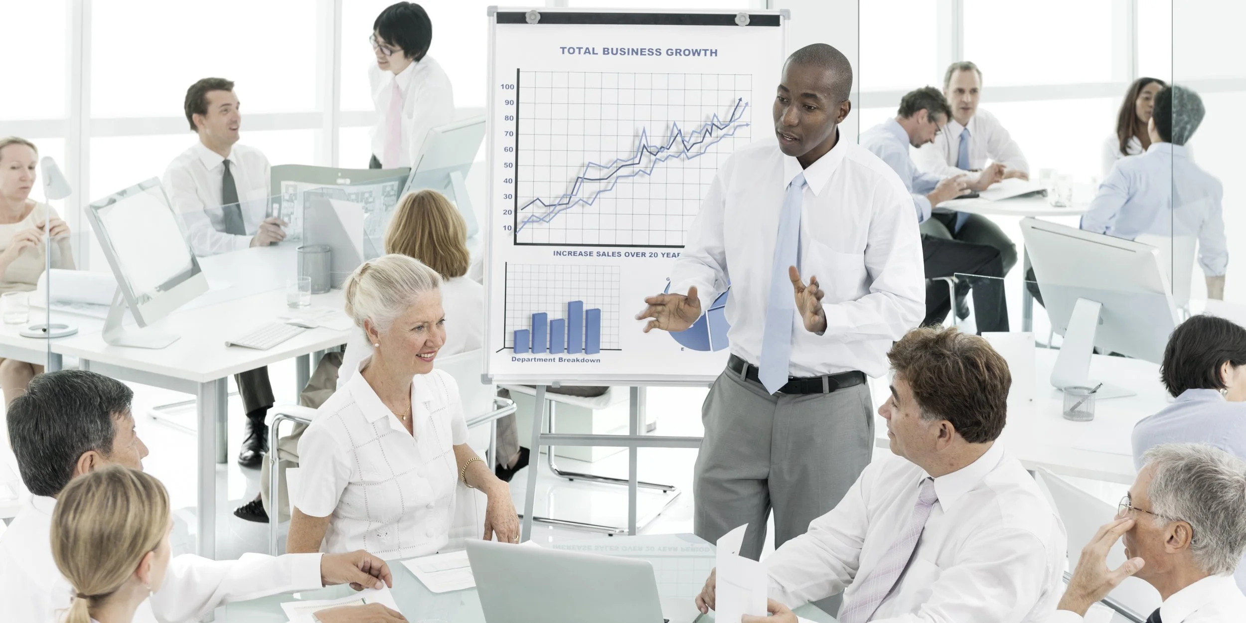 A business meeting or presentation in a modern office, with a man in a white shirt and light blue tie presenting data on a whiteboard, including charts showing business growth and sales increase over 20 years. Several people are seated around the room listening, taking notes, or engaging in the discussion.