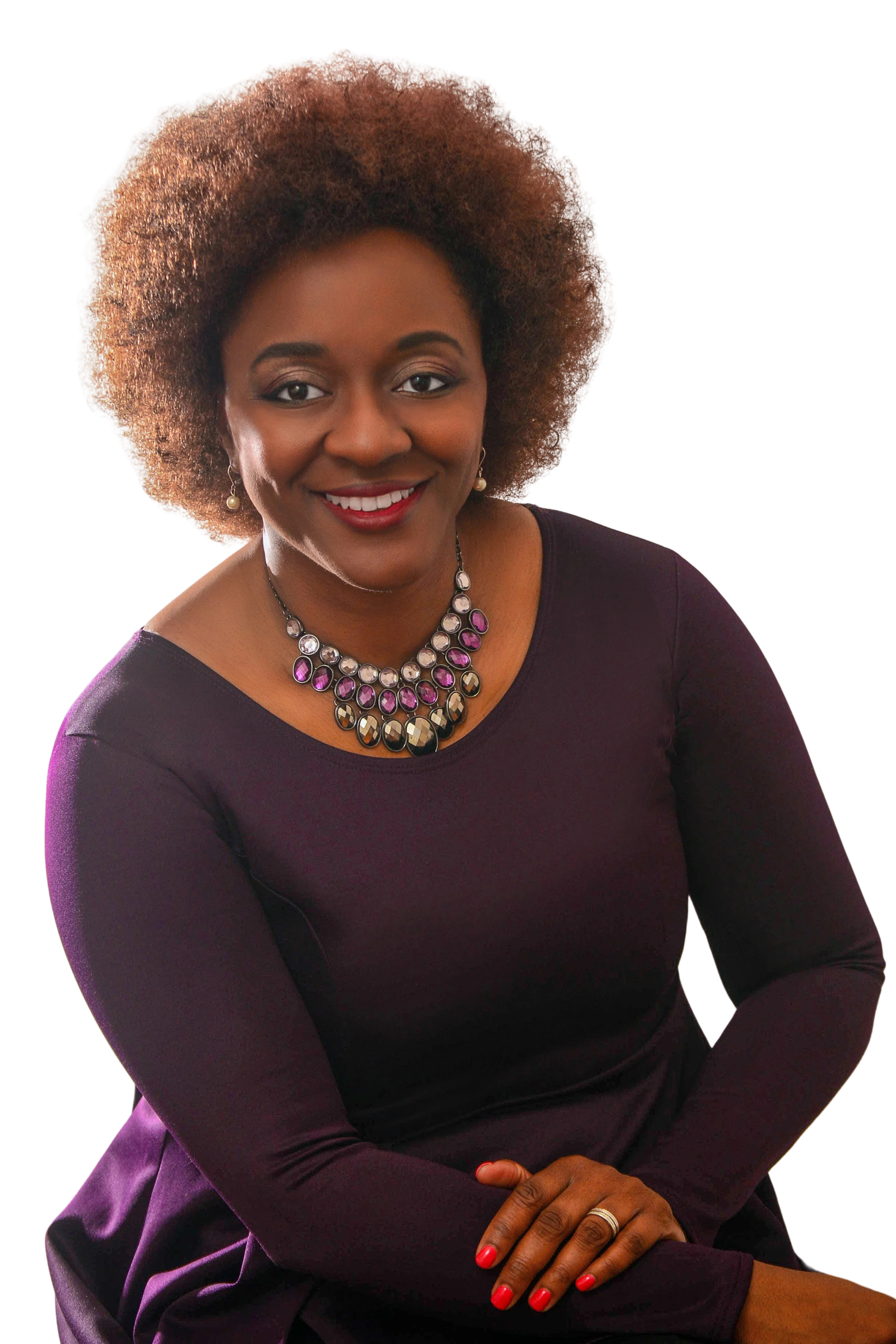 A smiling African American woman with a large brown afro hair, wearing a dark purple long-sleeve top, pearl earrings, and a statement necklace with pink and clear stones. She has red nail polish and a ring on her left hand, which is resting on her lap. The background is transparent.