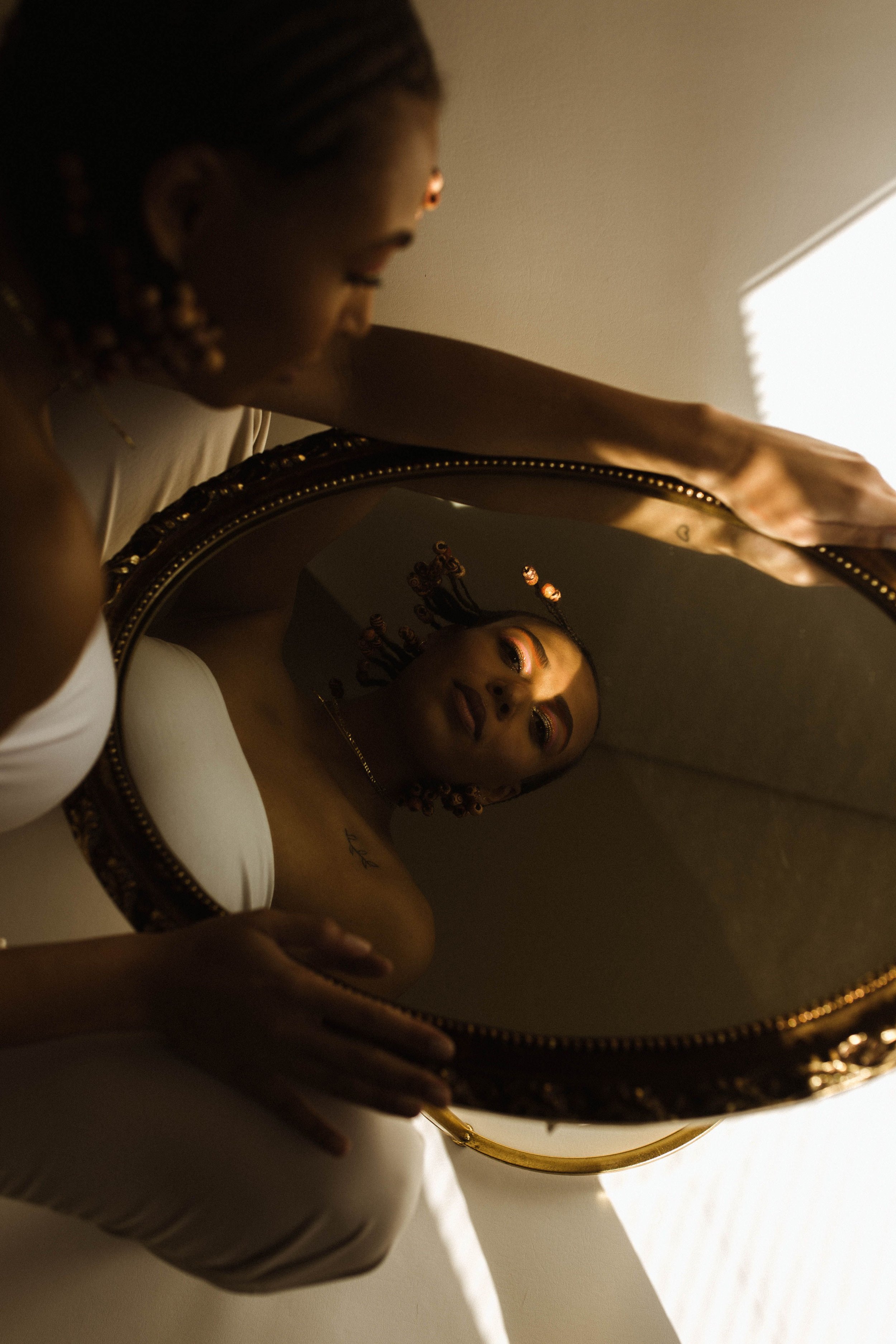 A woman looking at her reflection in a round mirror with a decorative gold frame, holding the mirror with her right hand. She has braided hair with beads and makeup with orange eyeshadow. The room has beige walls and soft lighting.
