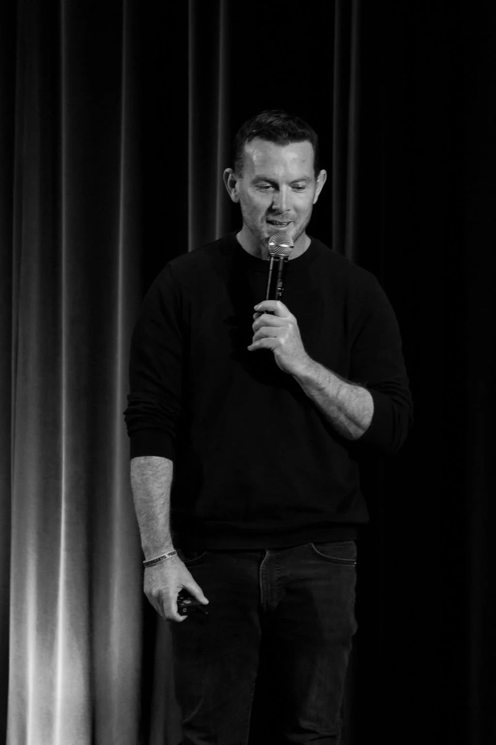 A man holding a microphone on stage with dark curtains in the background.