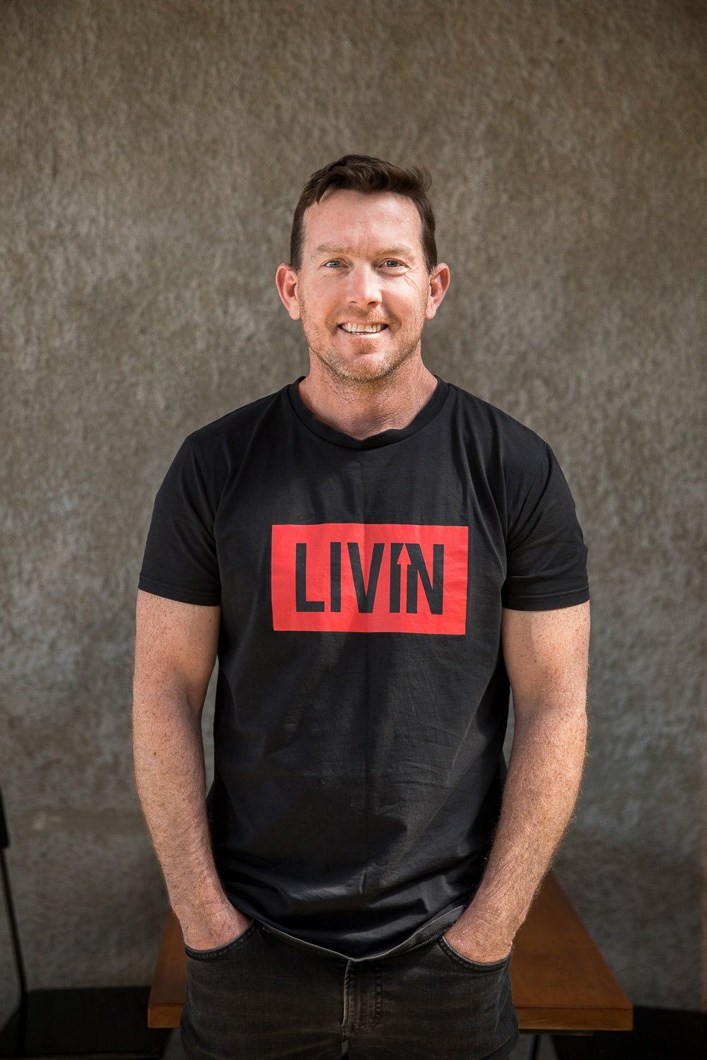 A man with short dark hair and a beard, wearing a black T-shirt with a red and black "LIVIN" logo, is standing and smiling in front of a textured gray background.