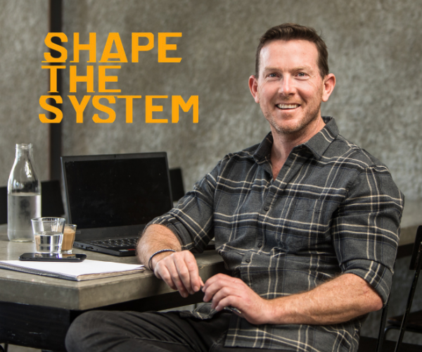 A smiling man in a plaid shirt sitting at a desk with a laptop, glass of water, and notebook, with the text 'SHAPE THE SYSTEM' in orange on a gray background.