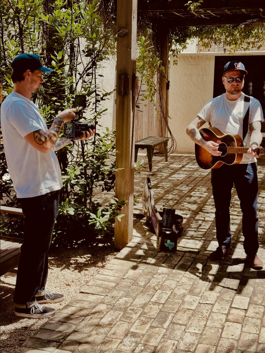 A man playing an acoustic guitar outdoors with a camera operator filming him, set in a shaded garden area with brick flooring and green foliage.