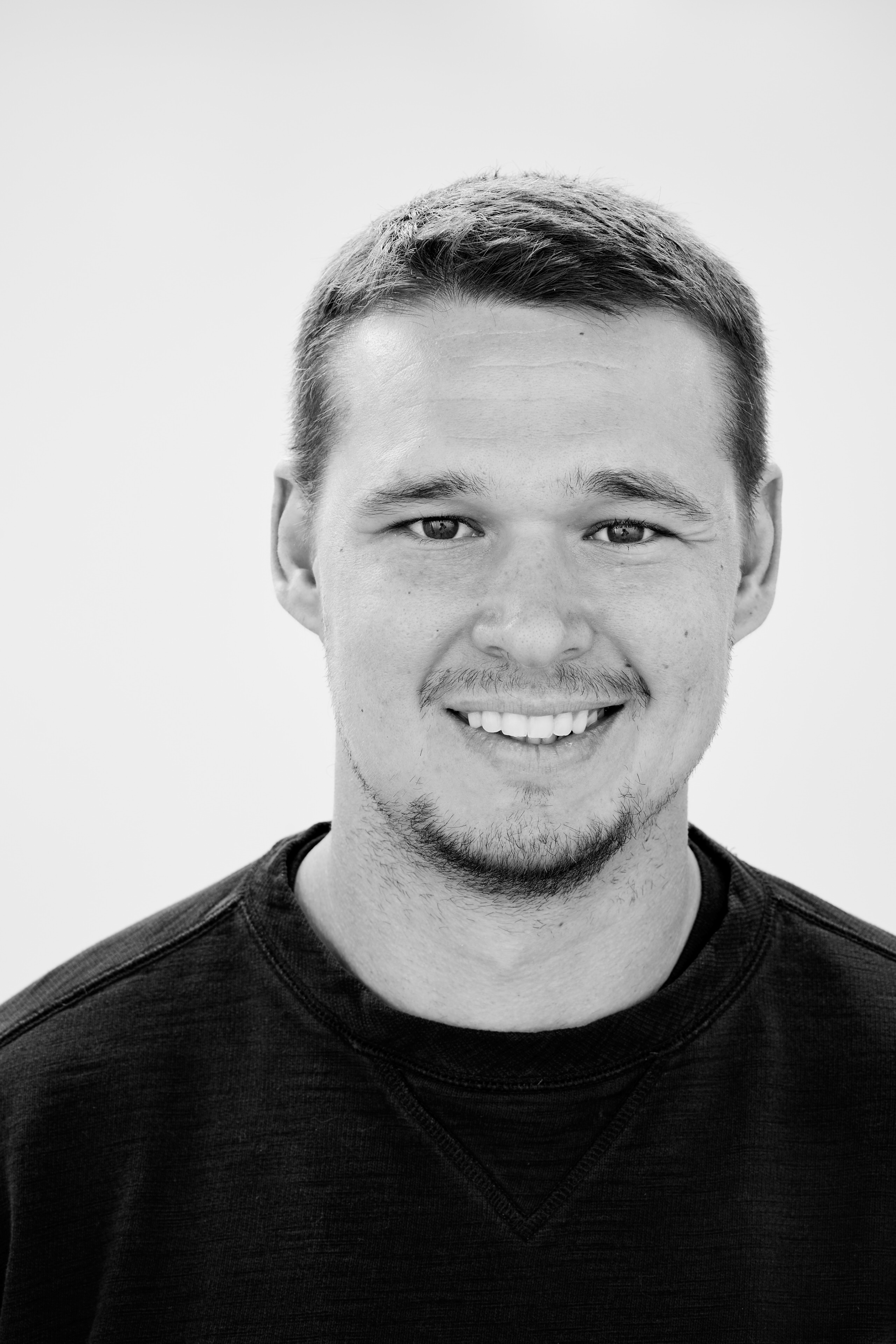 A black and white photo of a man smiling, with short hair, a mustache, and a goatee, wearing a dark shirt.