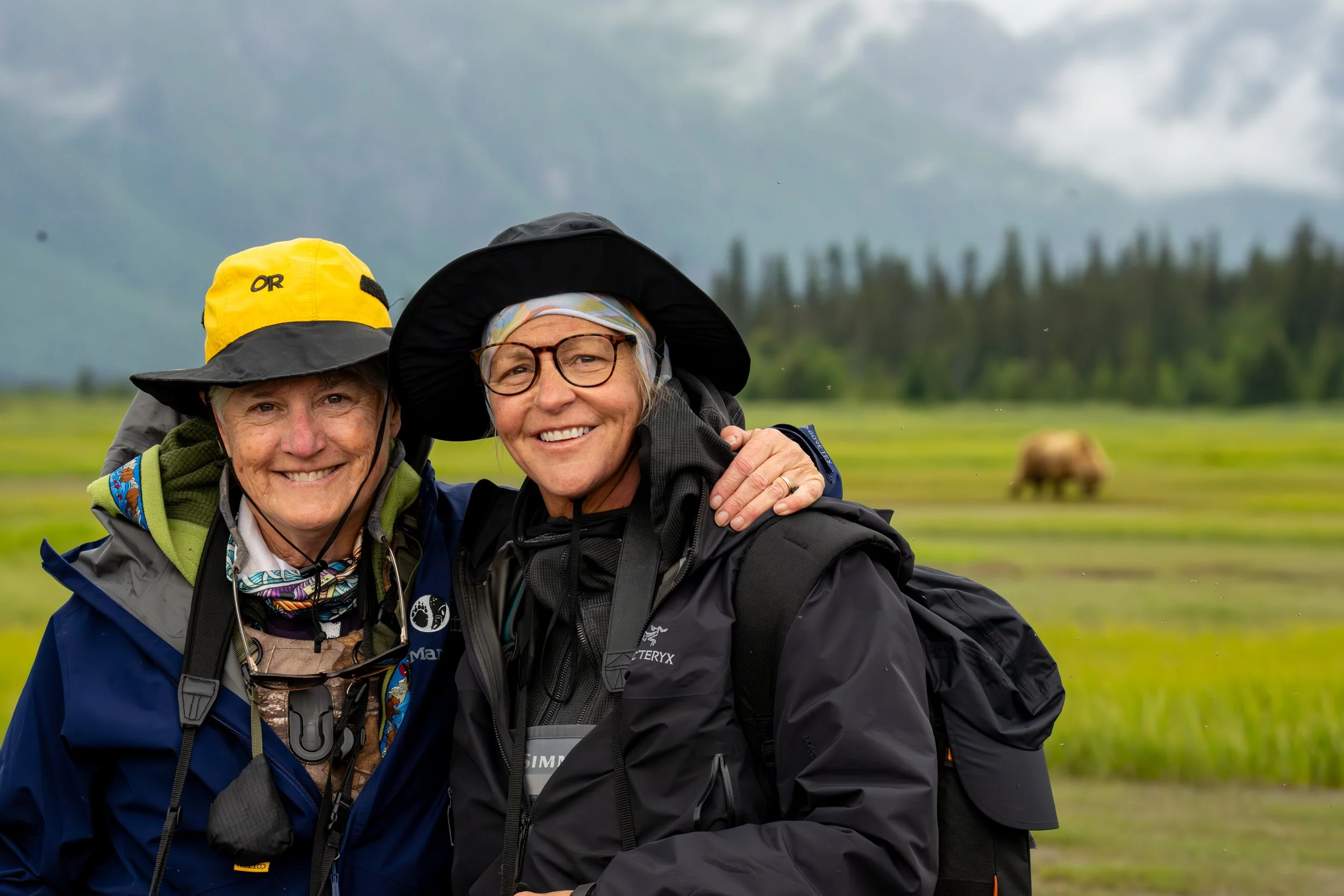 Two smiling women dressed in outdoor gear, standing close together with arms around each other, in a lush green landscape with mountains and a grazing cow in the background.