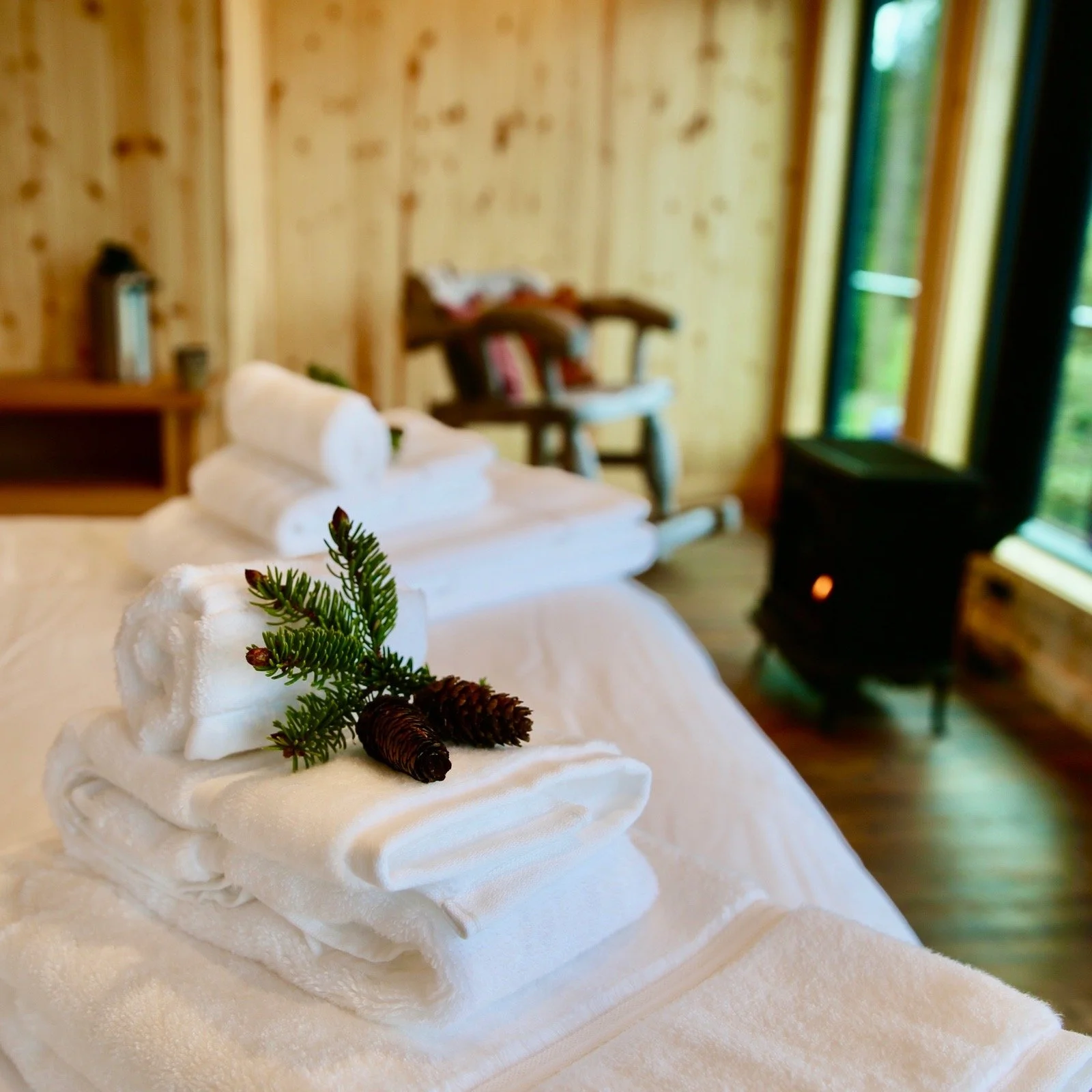 Stacked white towels on a bed with small pinecones and green pine branch for decoration in a cozy wooden room with green curtains and a heater.